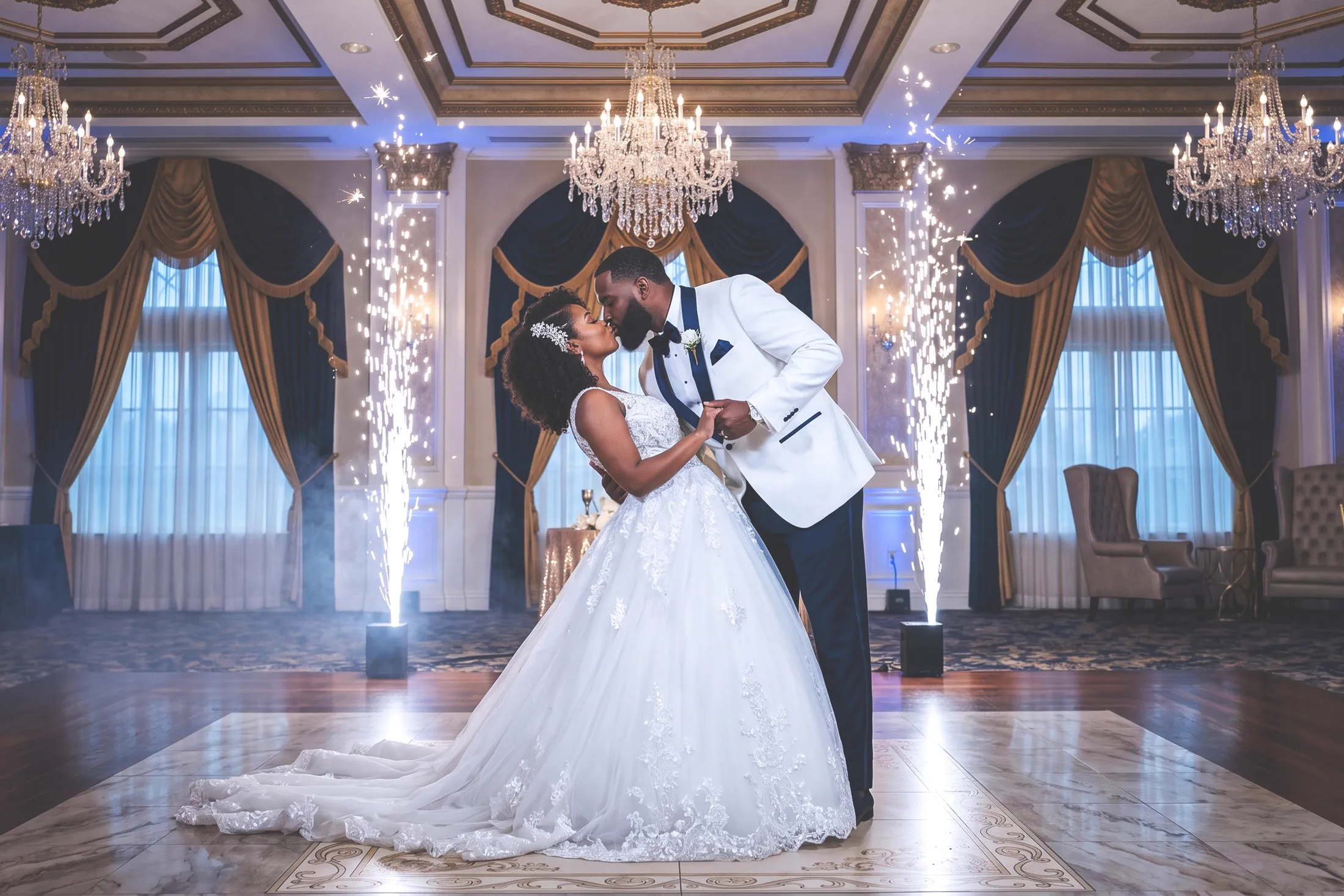 A newlywed couple kiss during a ballroom first dance framed by indoor sparklers.