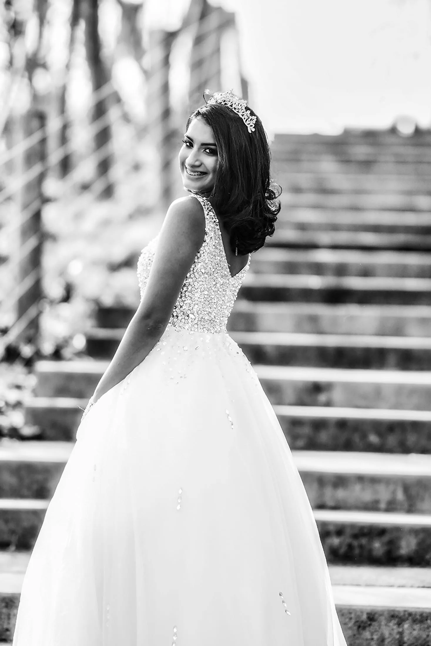A Sweet 16 celebrant in a sparkling gown smiles on a grand staircase.