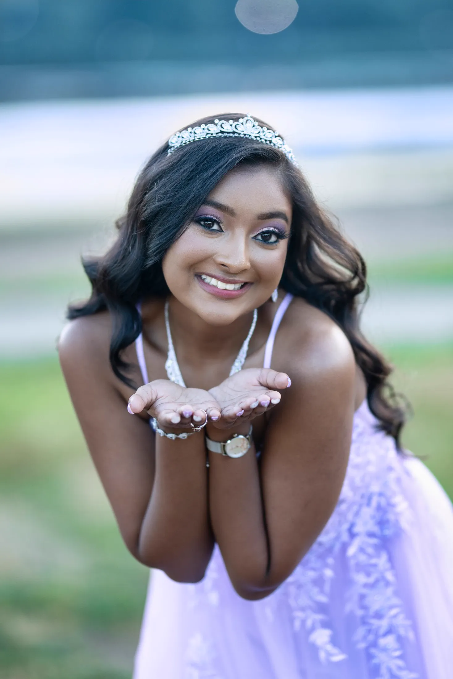 A Sweet 16 celebrant in a lavender gown and tiara smiles outdoors.