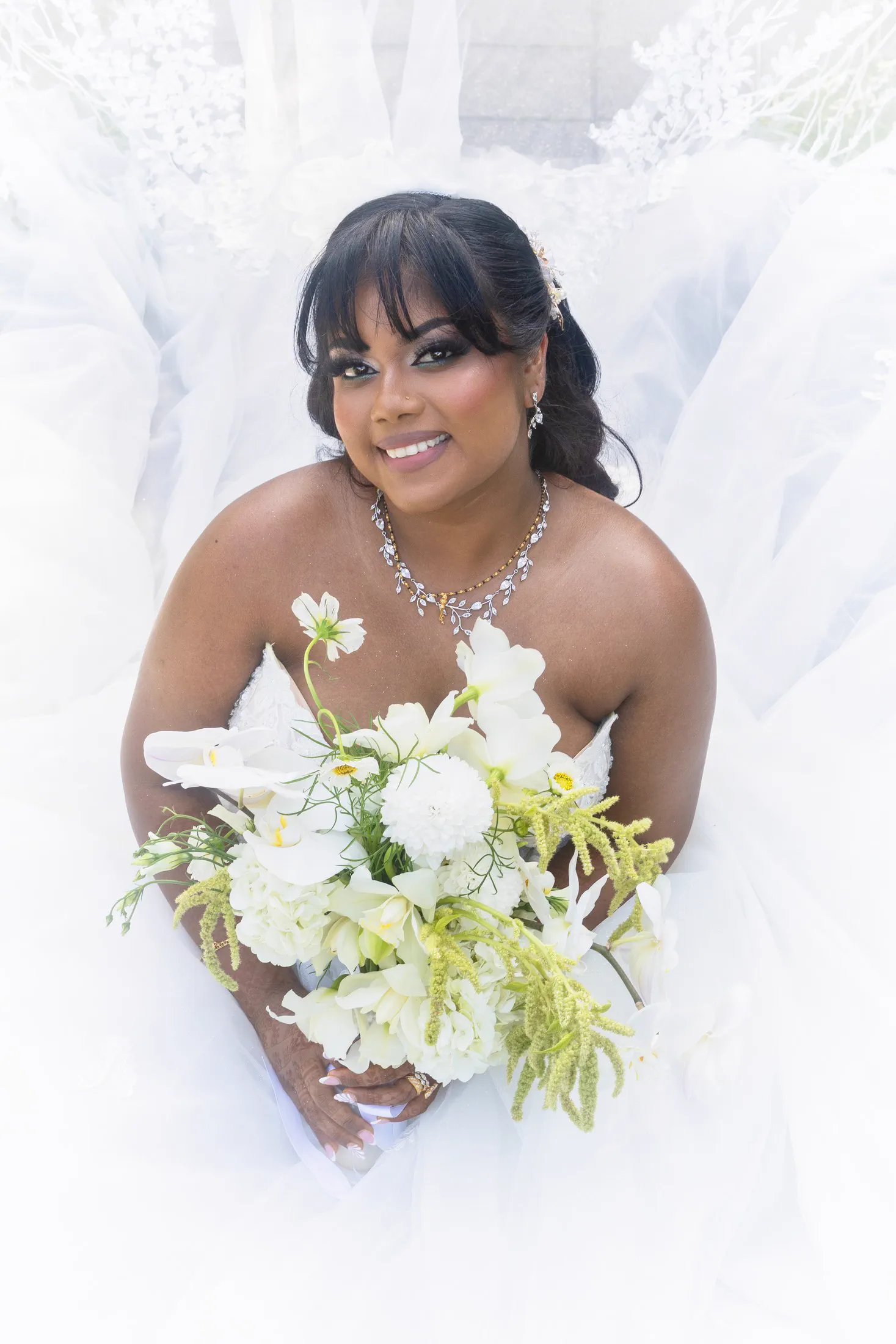 A bride sits beneath her veil holding a white and green bouquet.