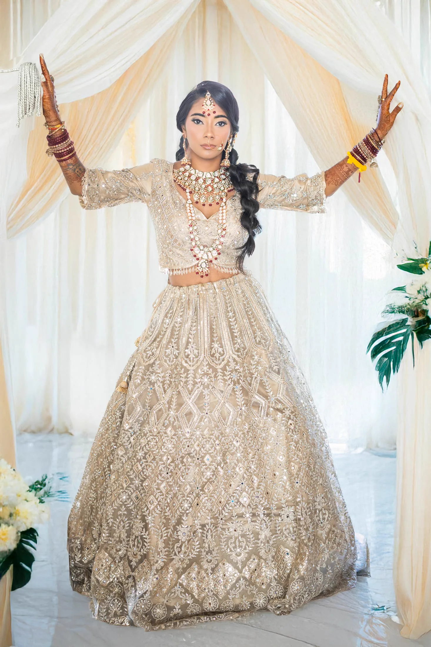 A bride raises her arms in a ceremonial pose, dressed in an embellished cream and gold lehenga.
