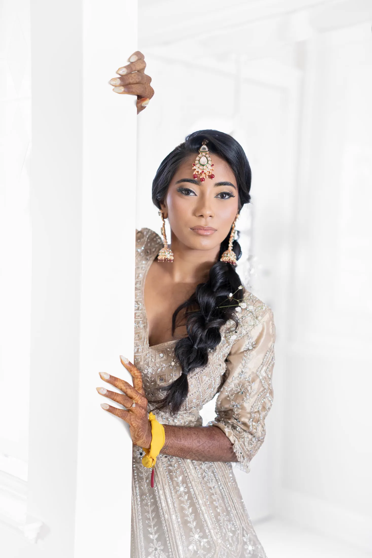 A bride in a beaded lehenga peeks around a white column, her hennaed hands on display.