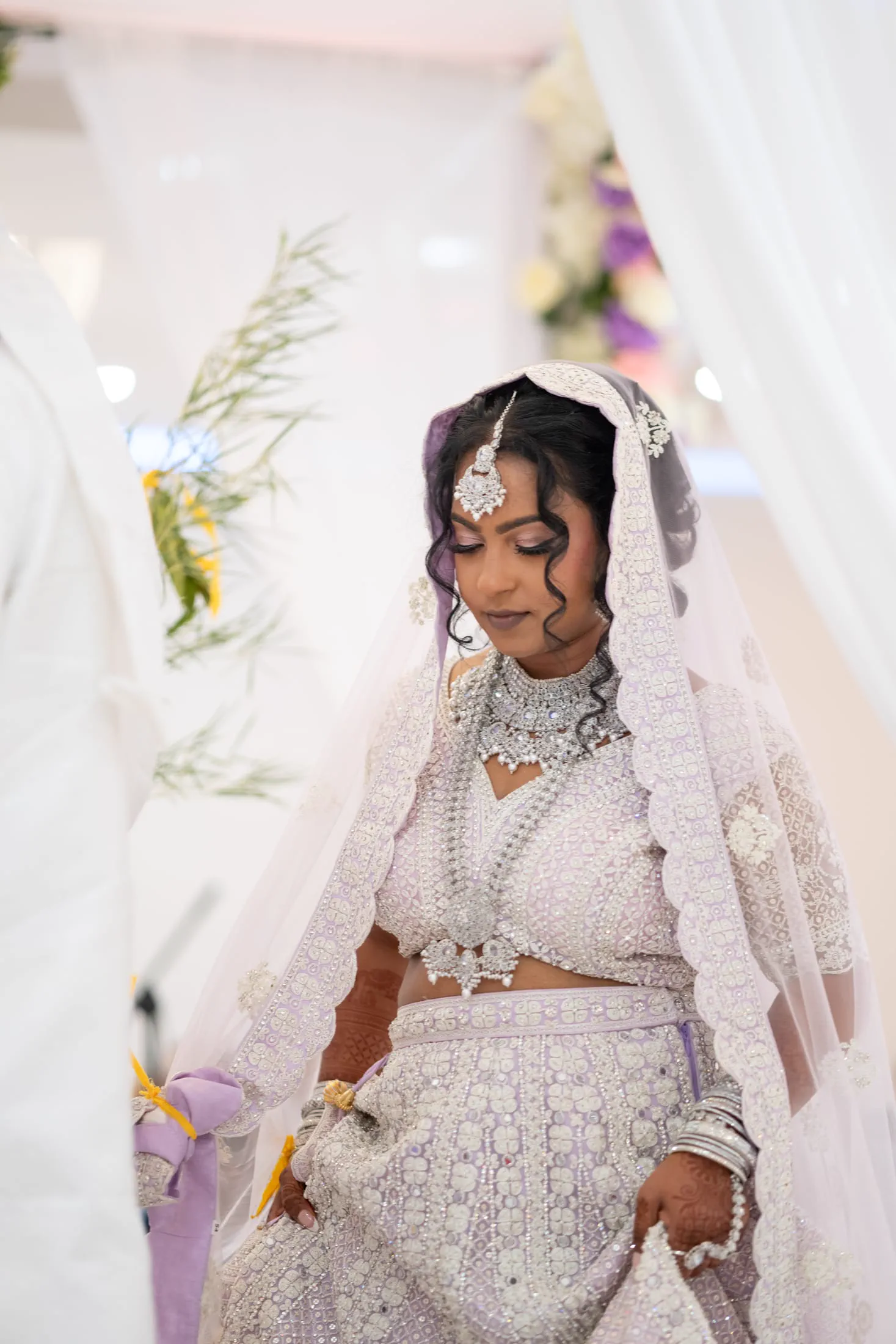 A bride in a purple and silver lehenga smiles beneath her veil.