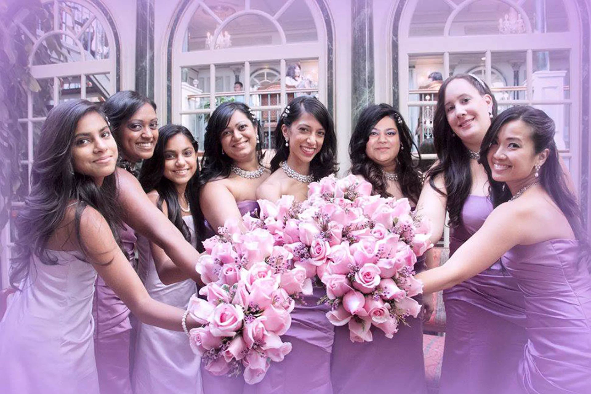 Bridesmaids in lavender gowns gather around the bride, holding pink rose bouquets together.