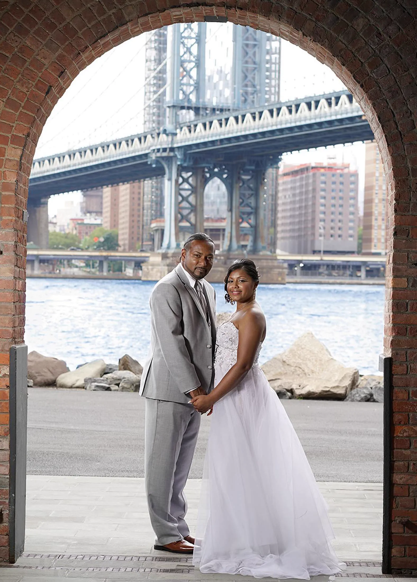 A couple stands framed by a brick archway beneath the Manhattan Bridge, the skyline and river behind them.
