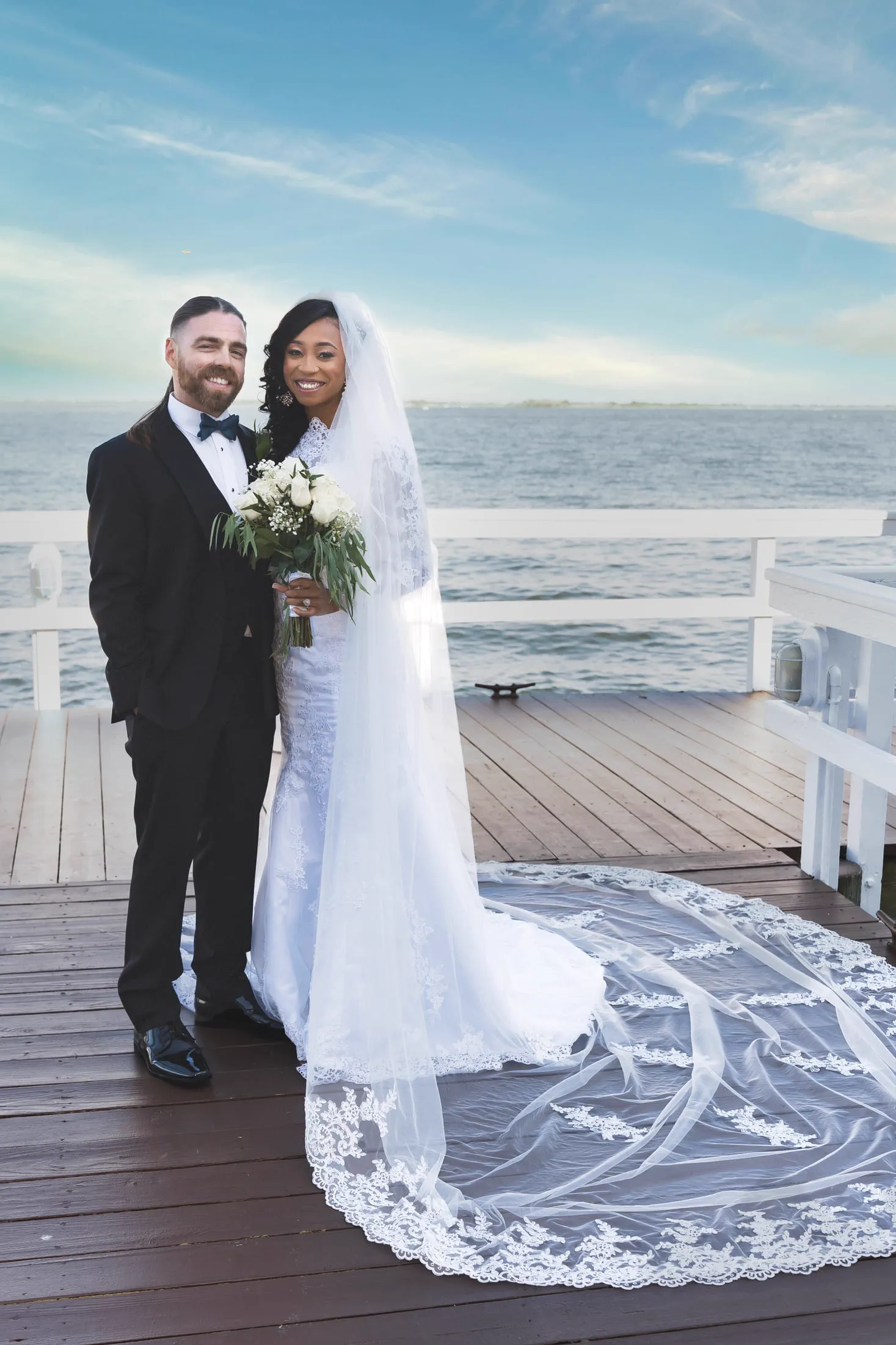 A bride and groom pose together on a waterfront dock, her veil spread across the boards.