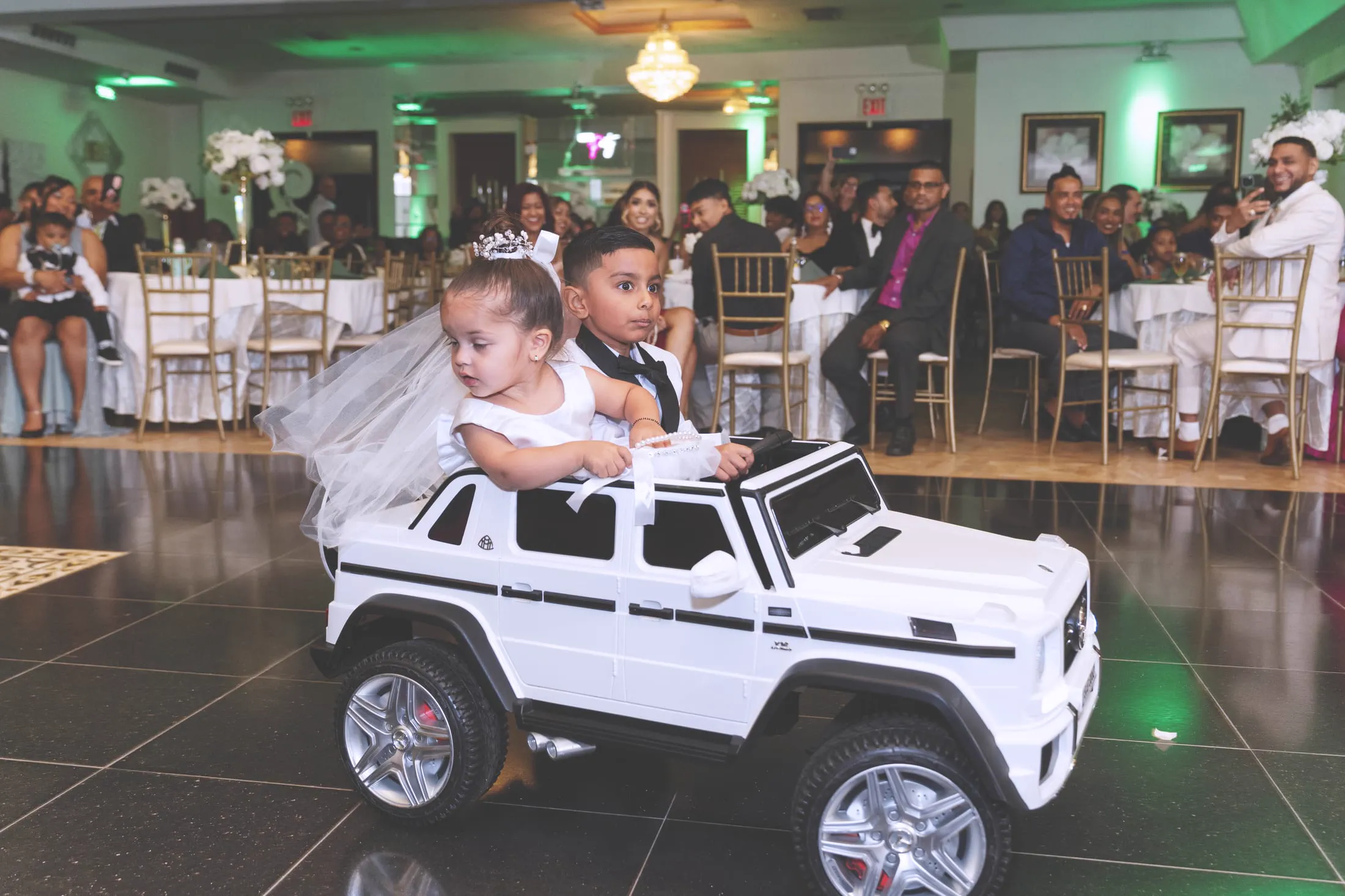 A flower girl and ring bearer ride a toy car at the reception.