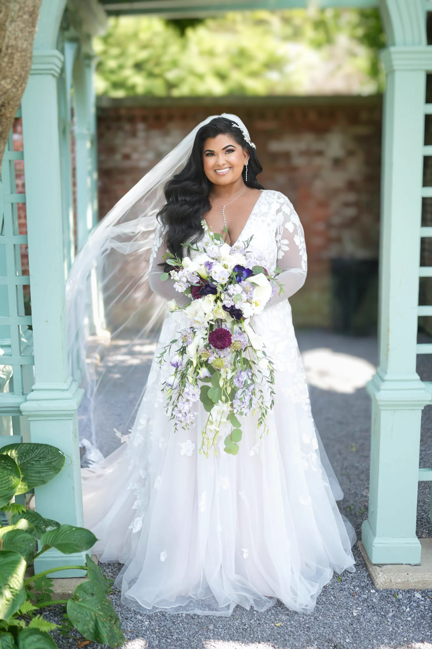 A bride beneath a garden gazebo holds a cascading purple and white bouquet.