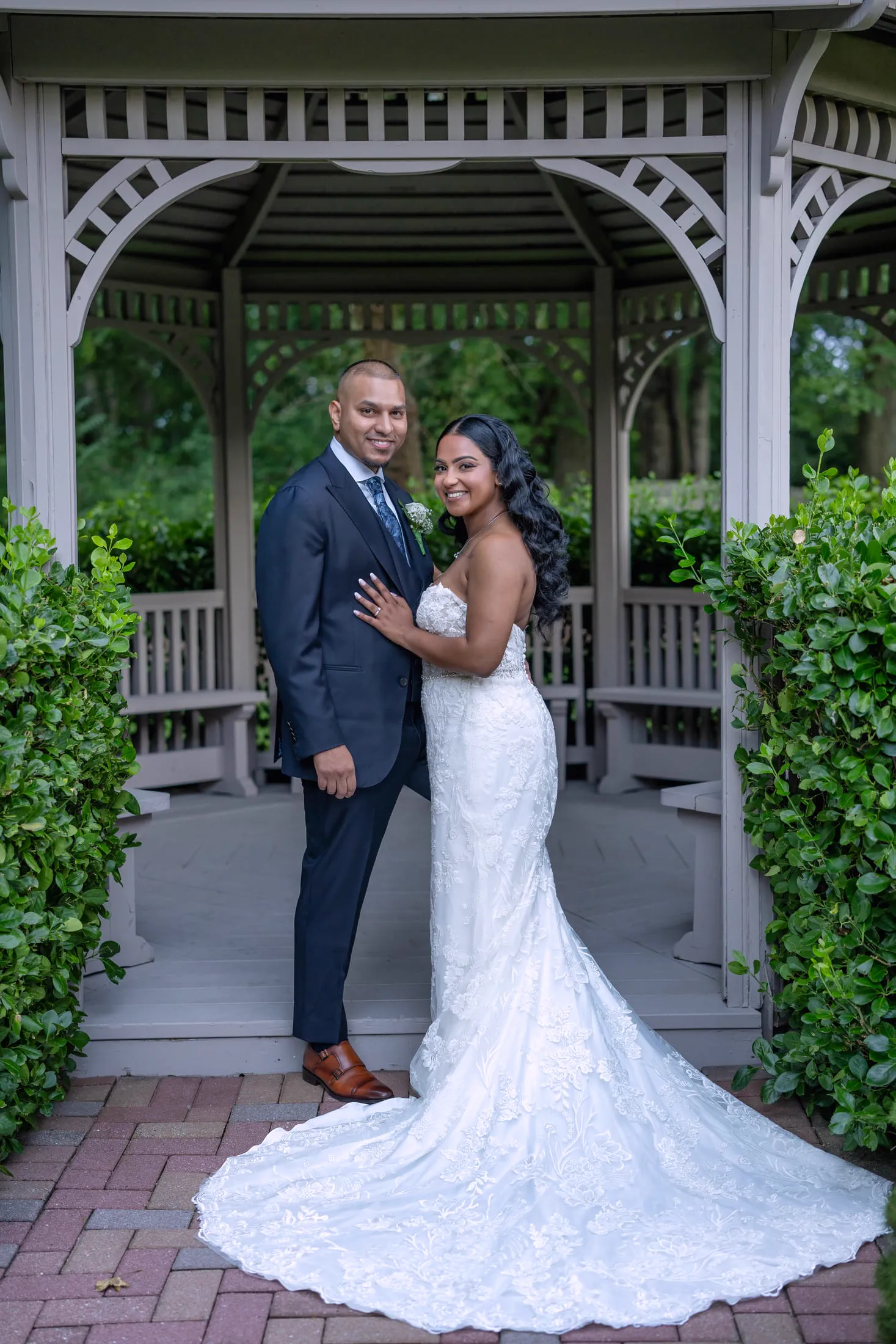 A groom in a navy suit stands with his bride beneath a garden gazebo.