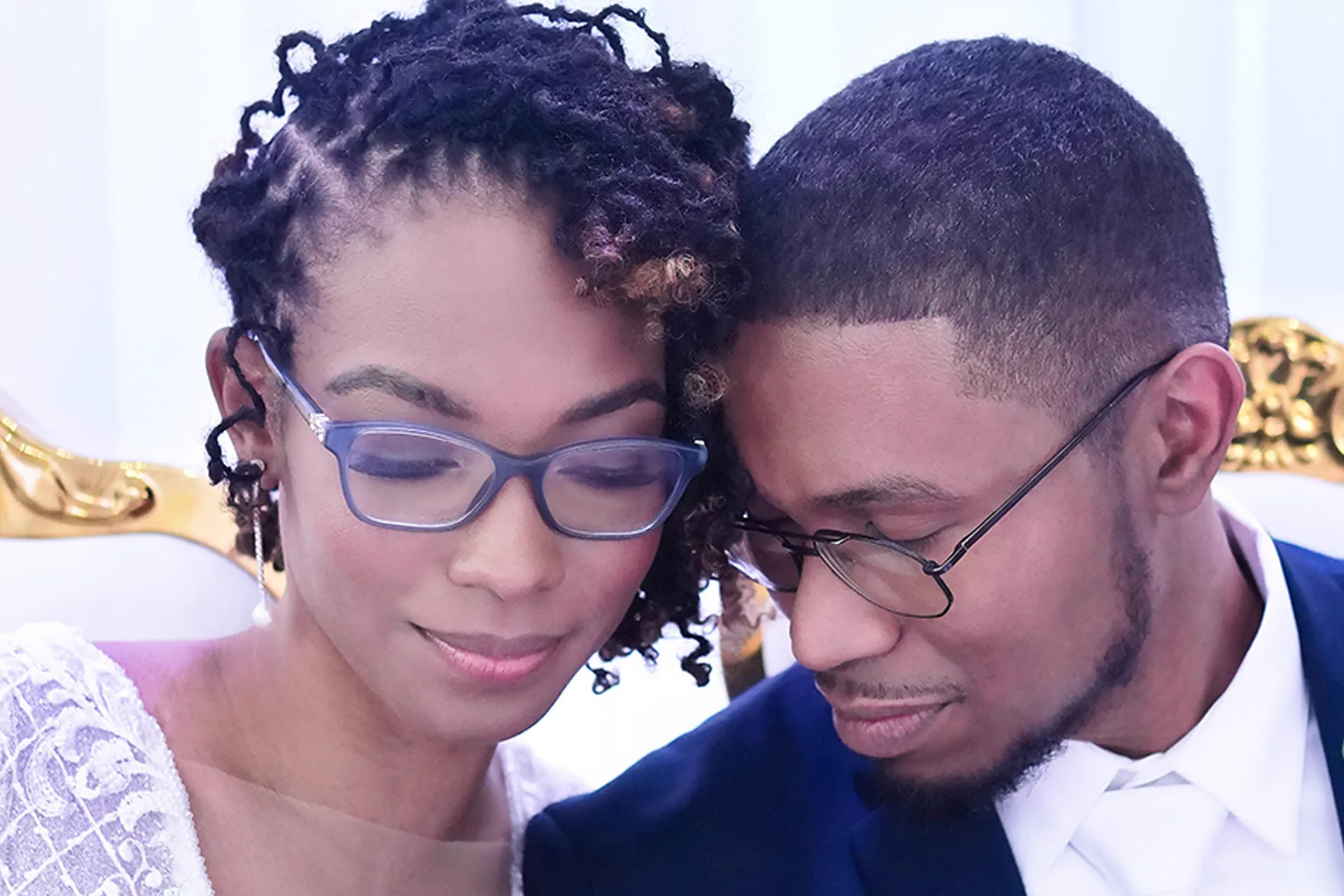 A couple, both wearing glasses, share a tender quiet moment forehead to forehead.