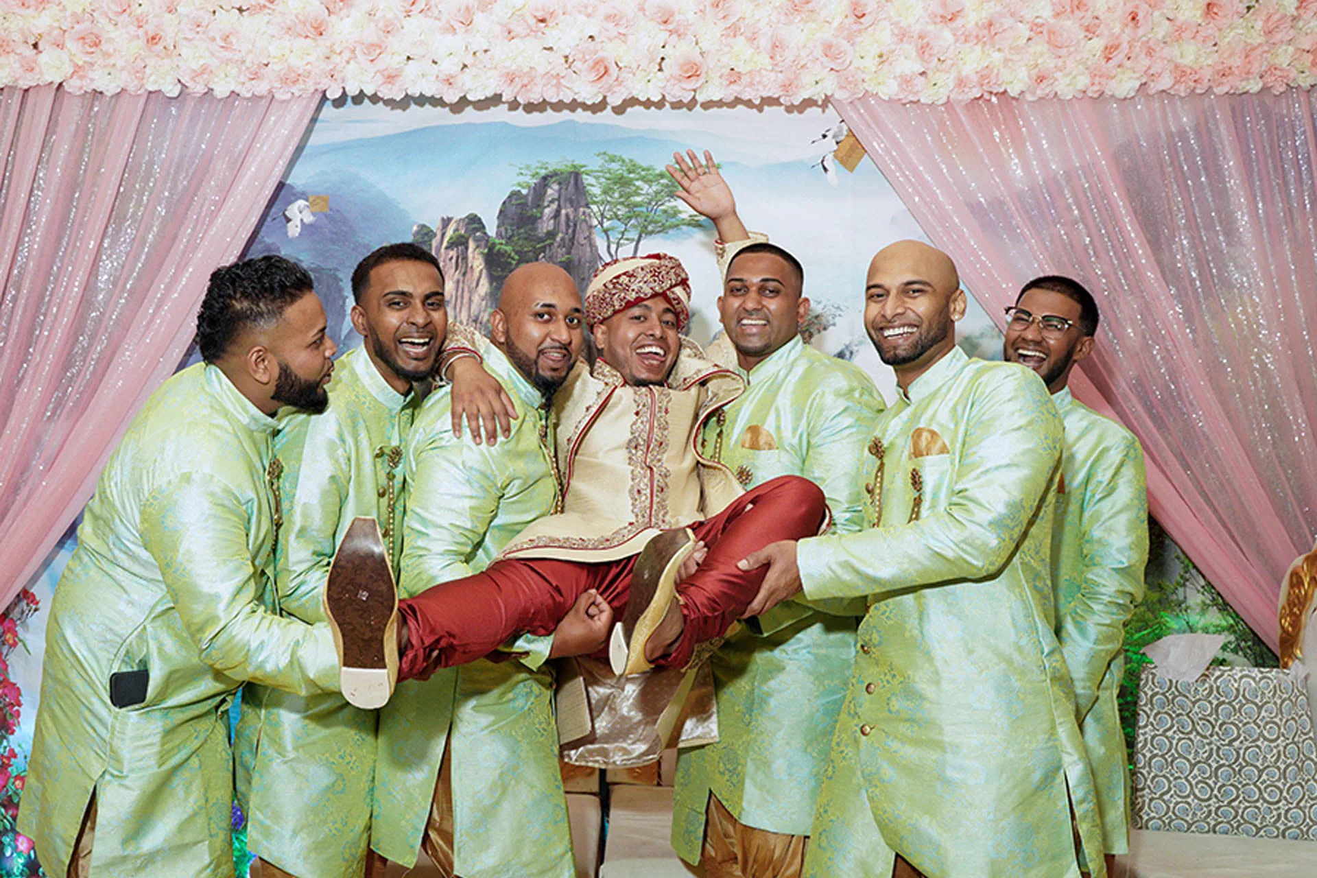 A groom in a red and cream sherwani is joyfully lifted by his groomsmen.