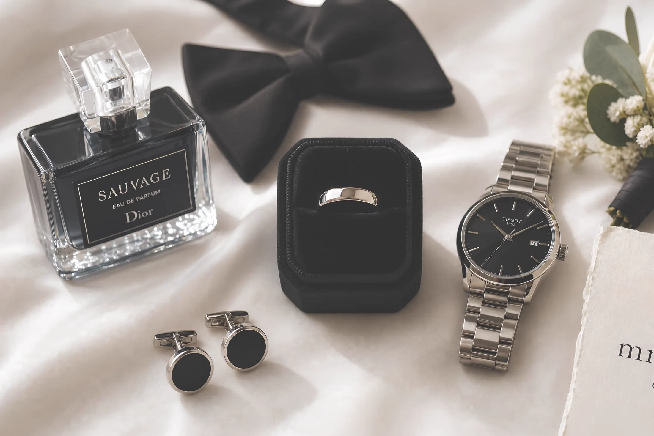 A flat lay of the groom's wedding day details: cologne, bow tie, ring, watch, and cufflinks.