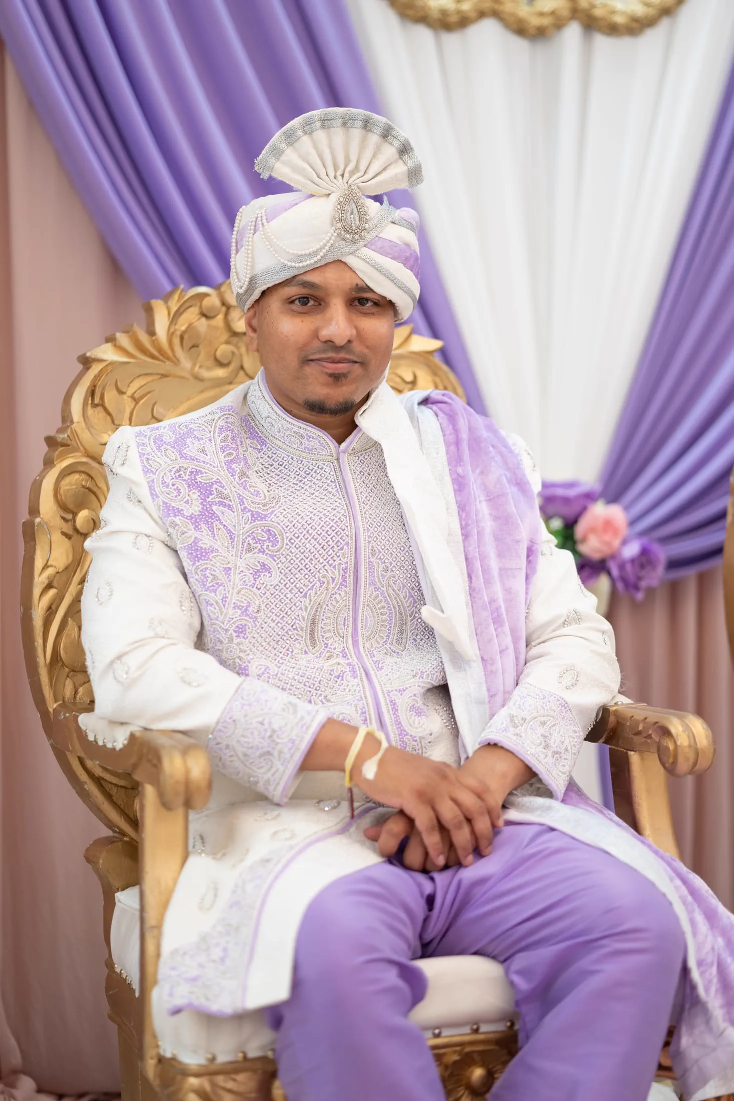 A groom in a purple and gold sherwani and turban sits on a ceremonial throne.