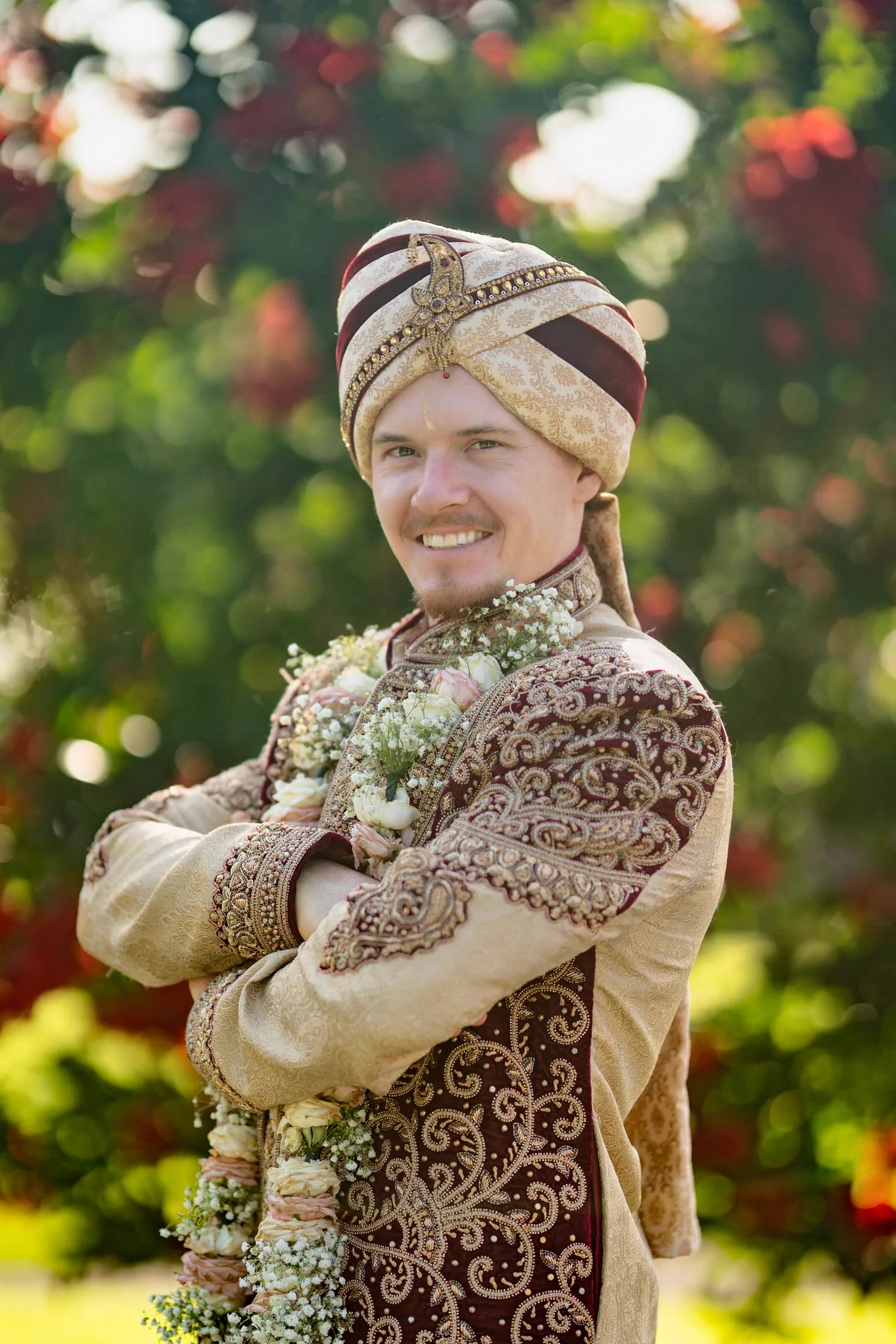 A groom in a traditional turban and floral garland smiles among garden roses.