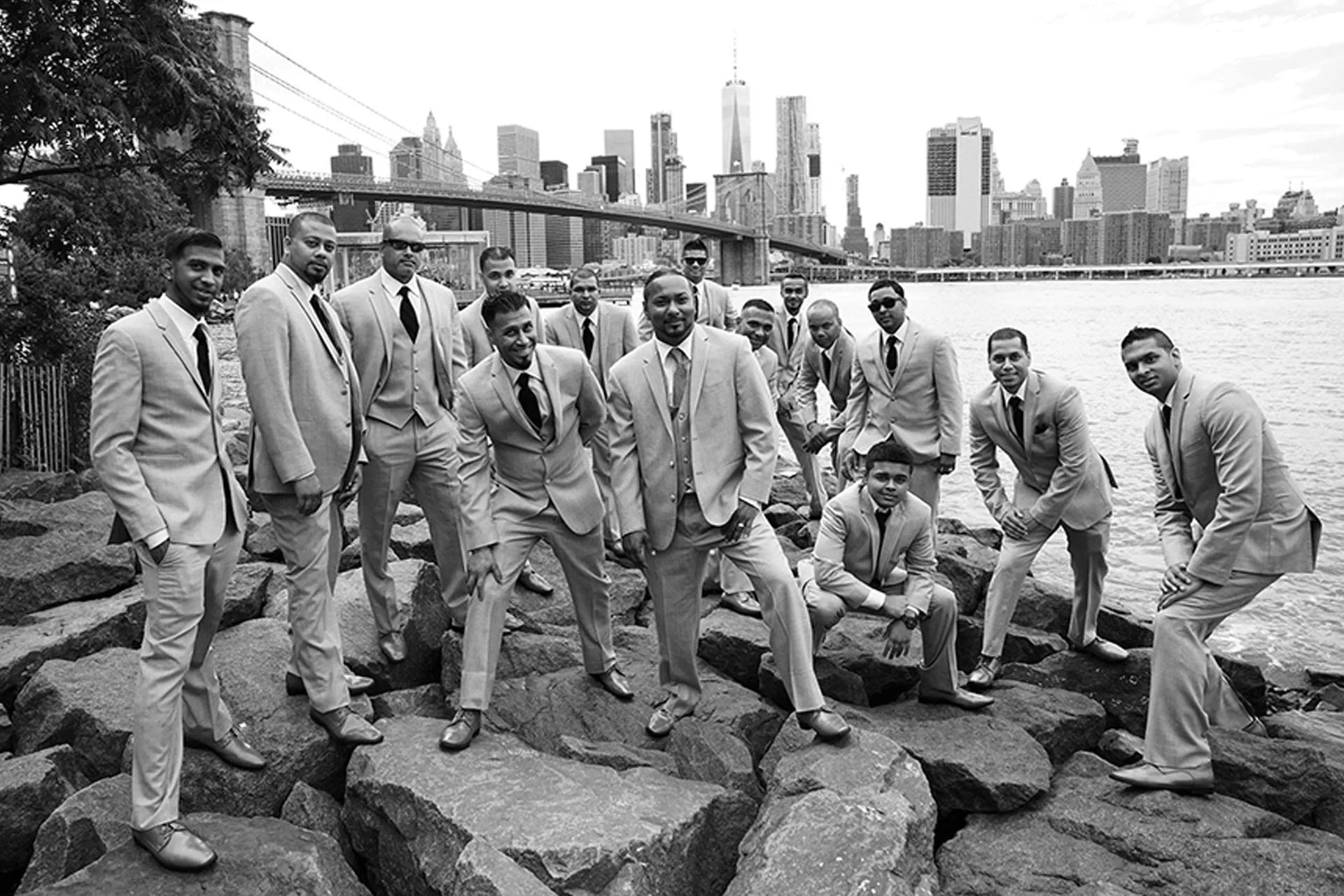 A groom and his groomsmen pose on rocks along the East River with the Brooklyn Bridge and Manhattan skyline behind them.