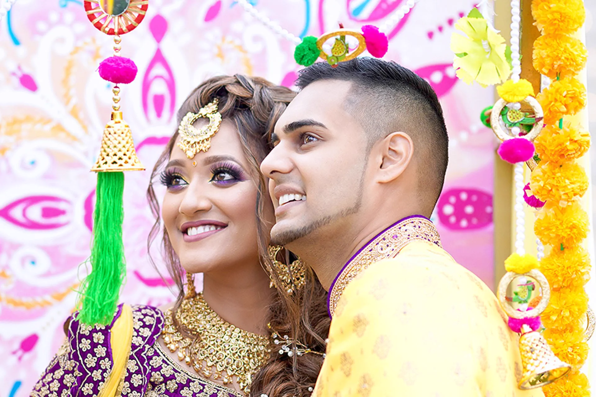 A couple leans close together beneath colorful Mehndi decorations.