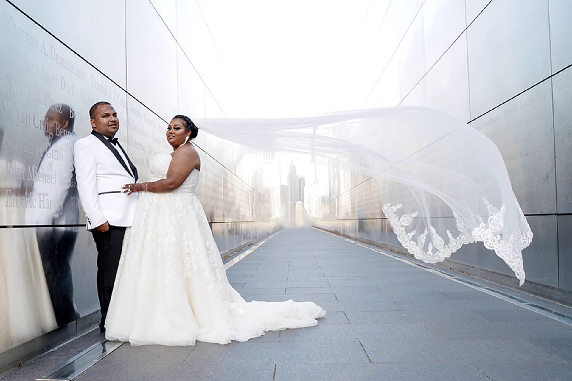 A newlywed couple pose at the Empty Sky memorial as the bride's veil sweeps across the walkway, the New York City skyline behind them.