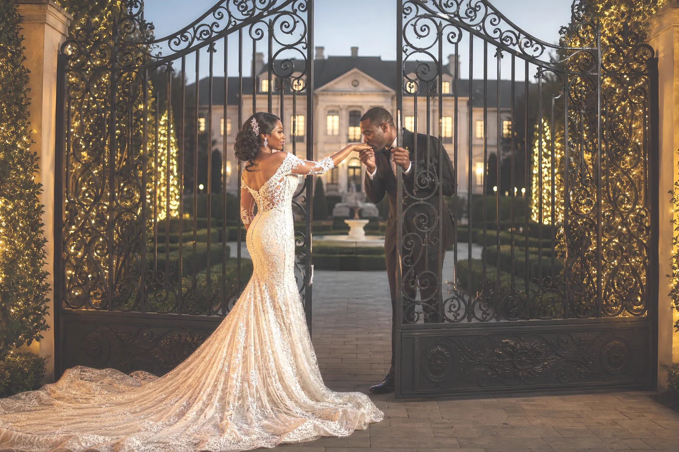 A bride and groom meet between ornate estate gates at golden hour.