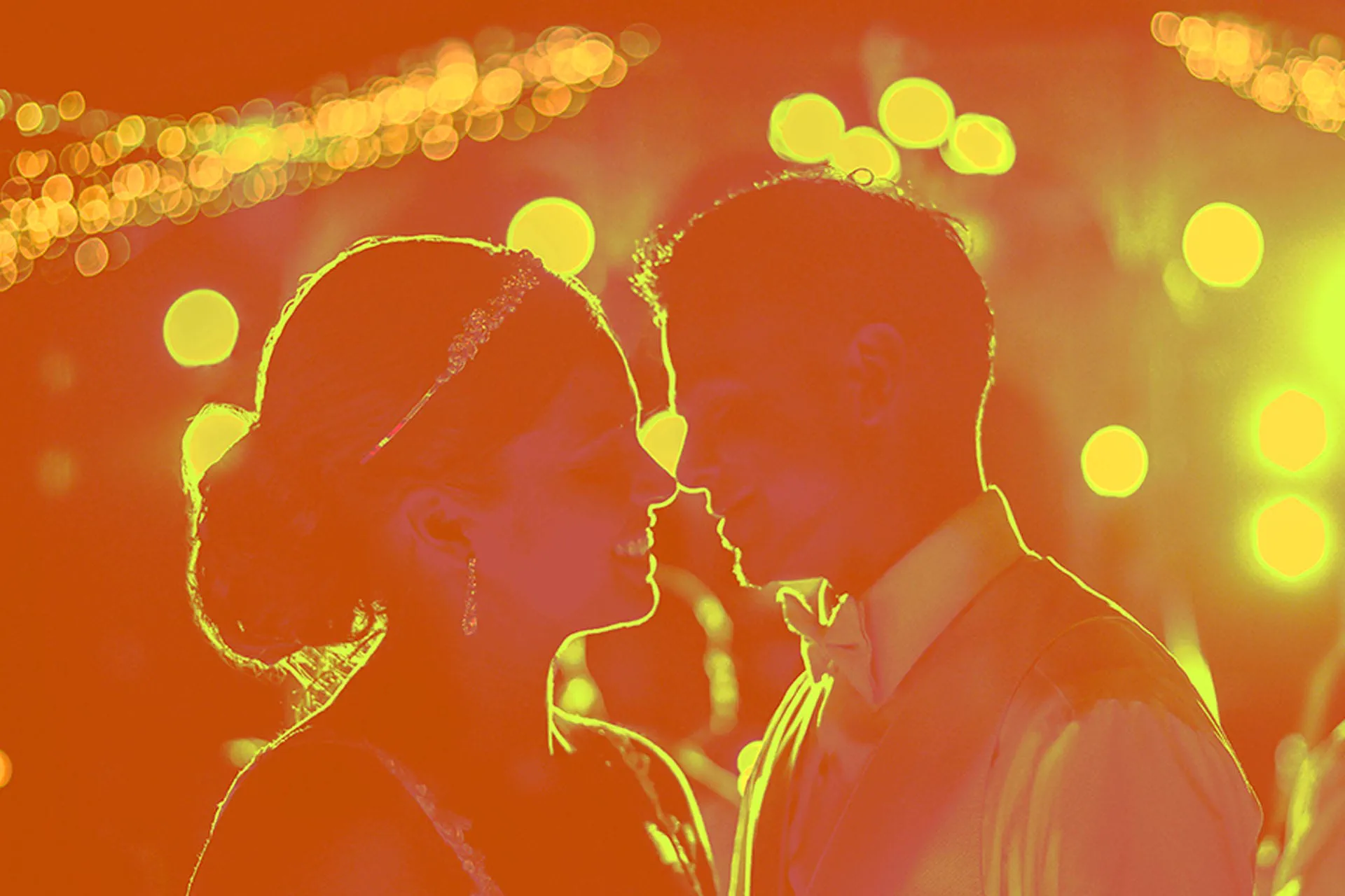 A couple's silhouette almost meets in a kiss against warm string lights.