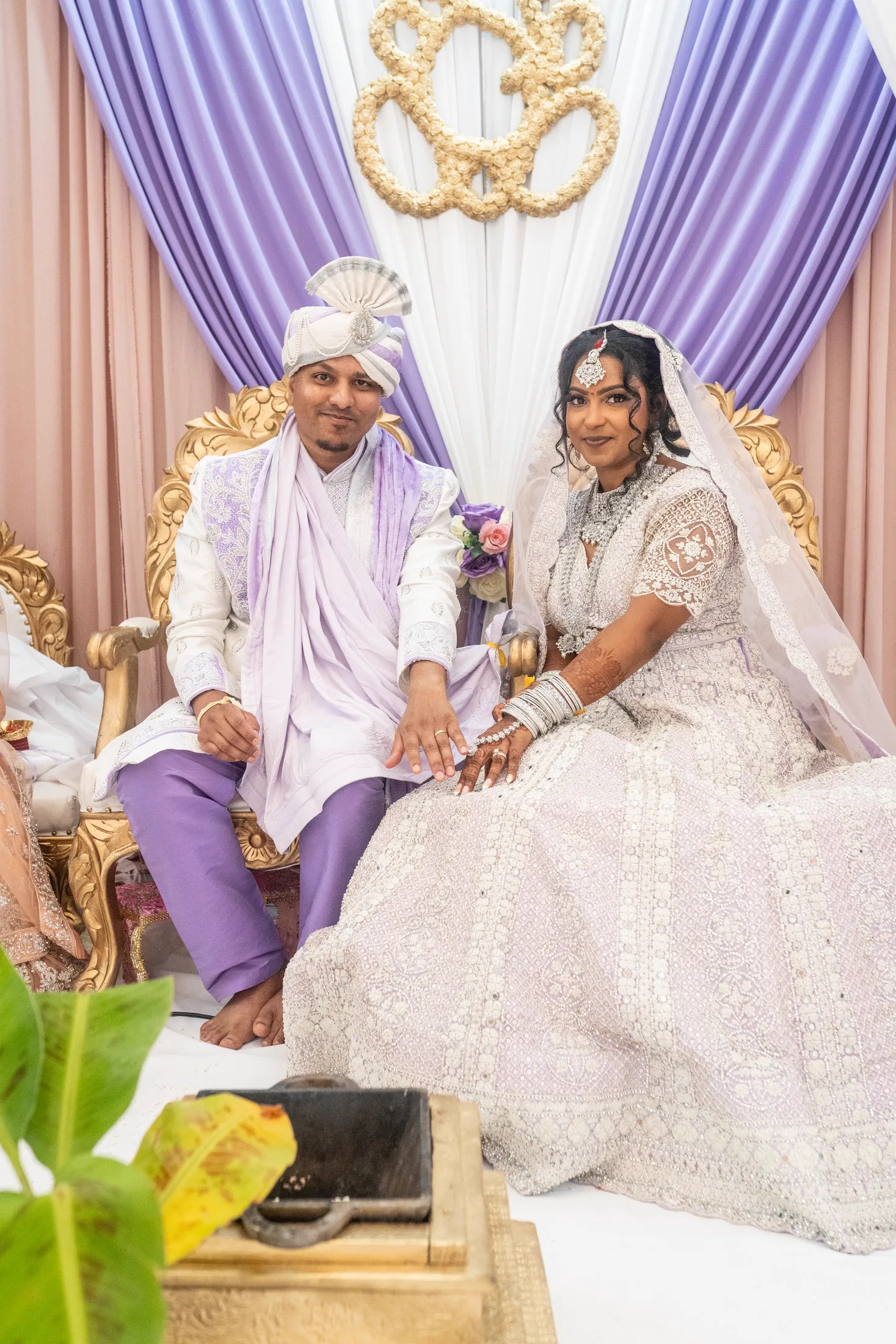 A South Asian newlywed couple sit together on an ornate wedding stage.