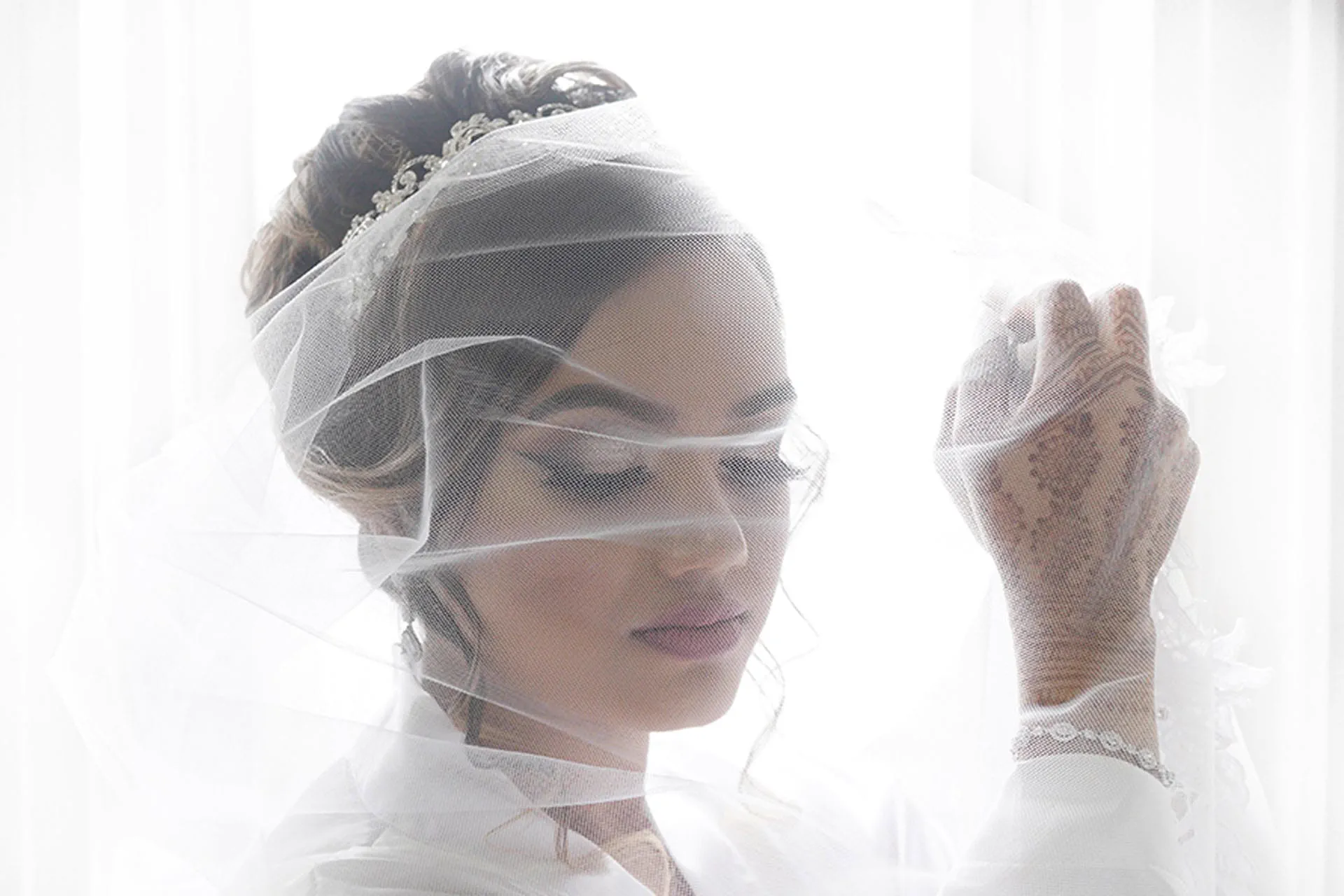 A bride wrapped in her veil against a bright window, her hennaed hand raised.