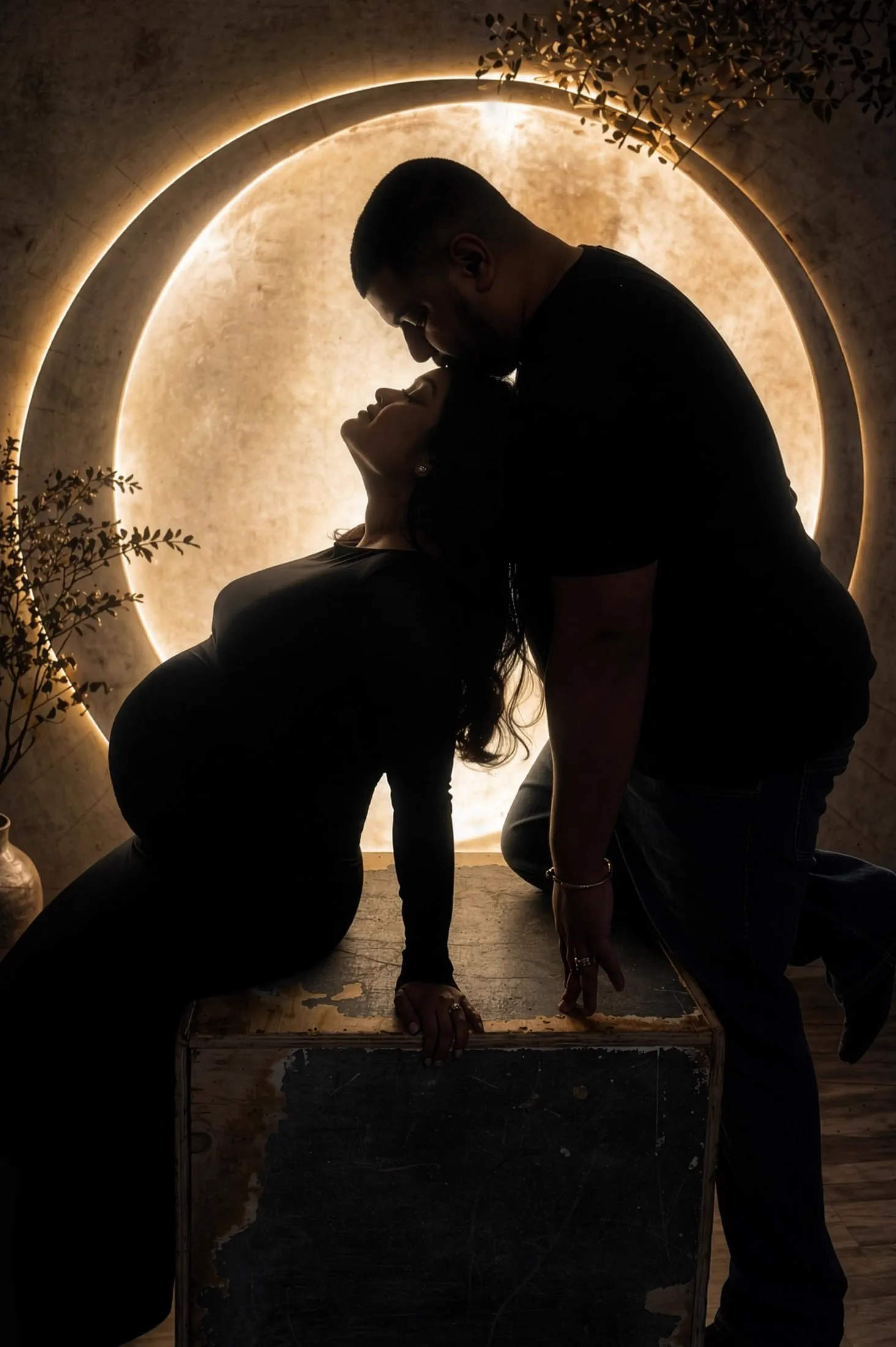 An expecting couple in silhouette share a tender embrace before a glowing circular light.