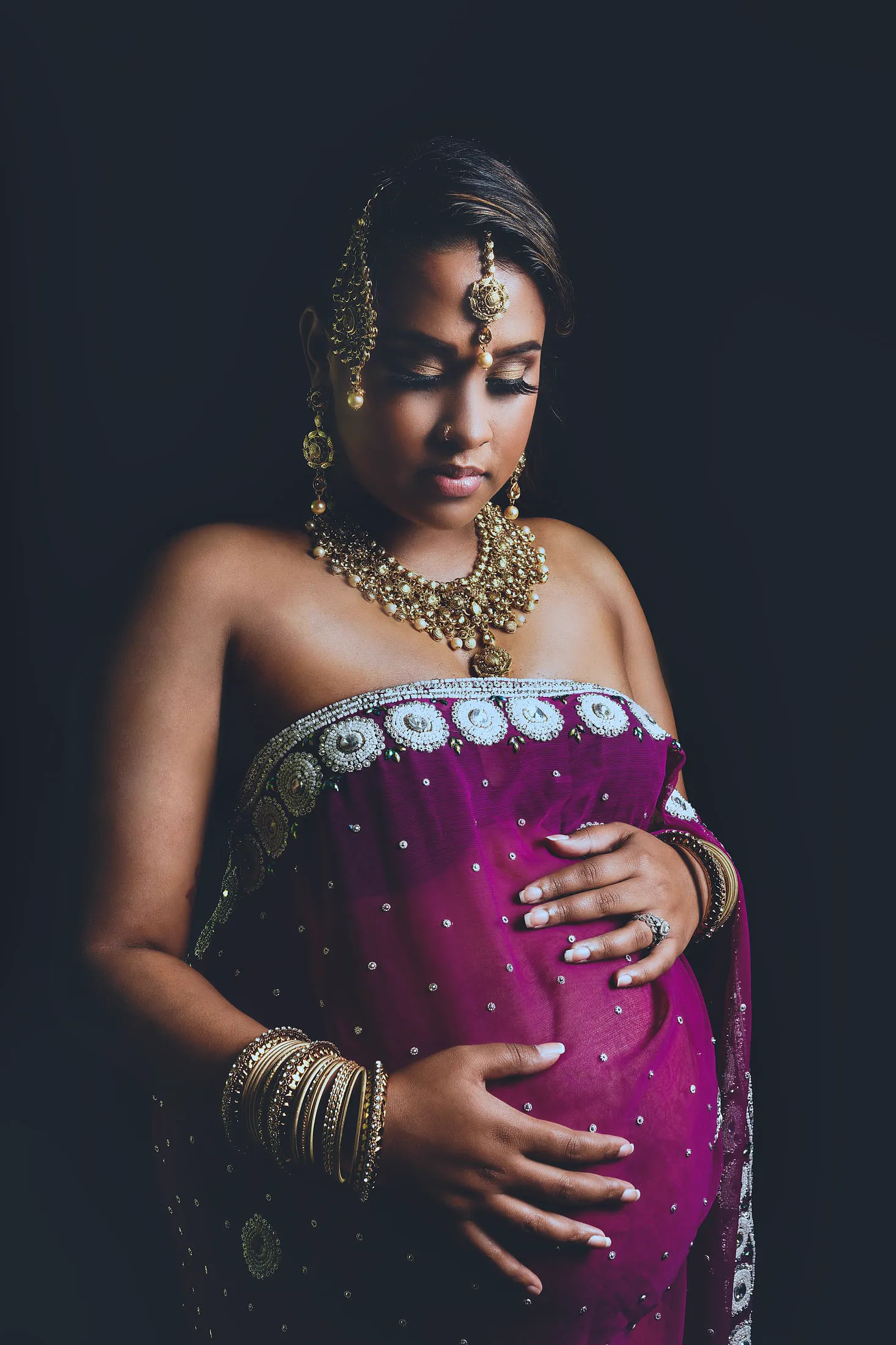 An expecting mother in a magenta sari and gold jewelry cradles her belly.