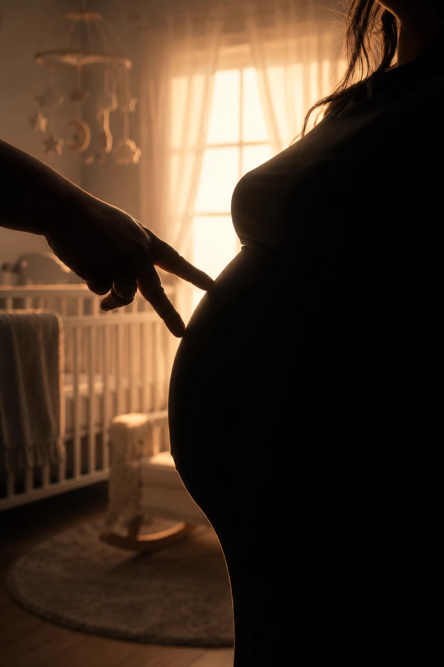 A silhouetted hand rests on an expecting mother's belly within a softly lit nursery.