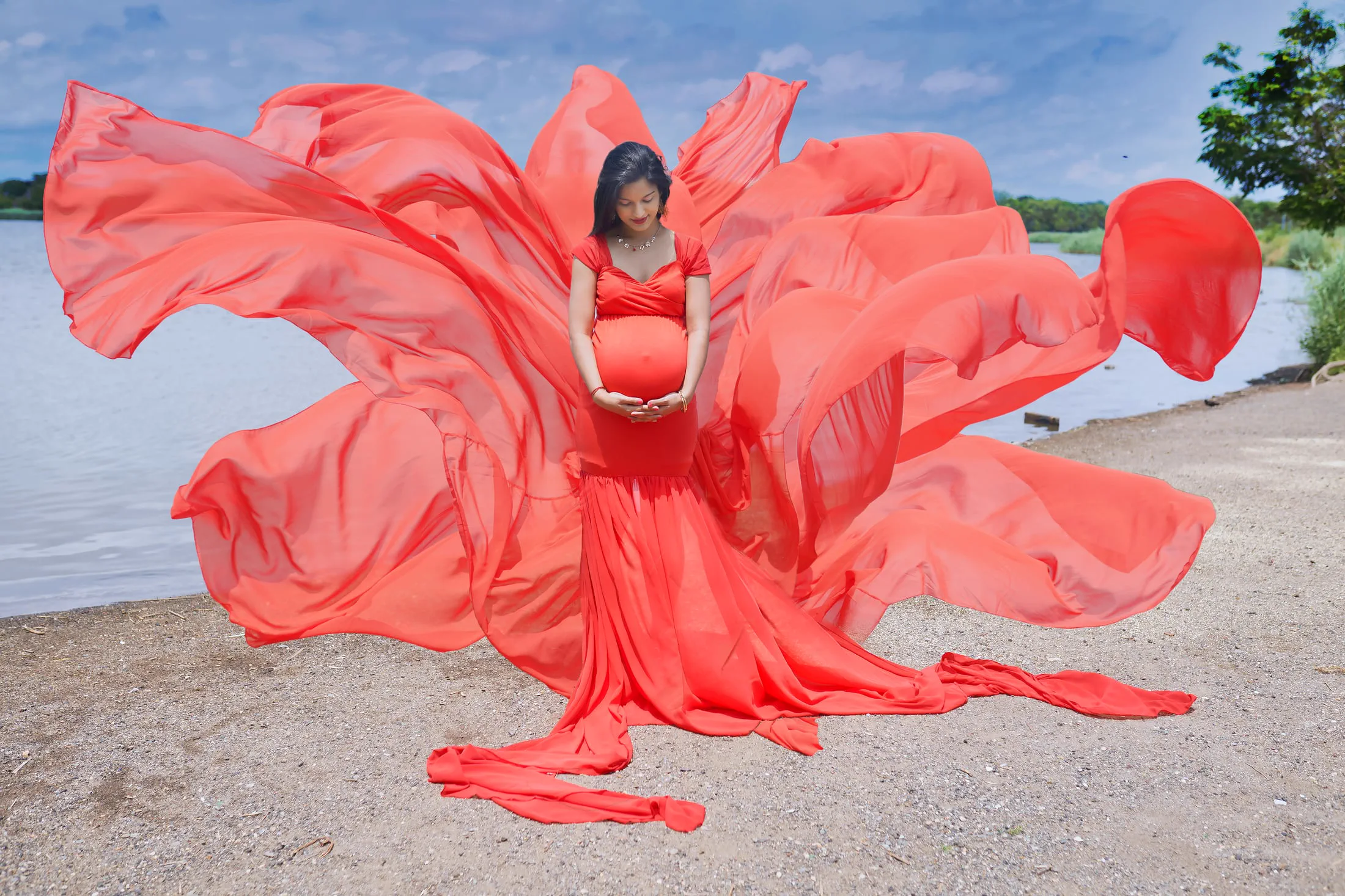 An expecting mother in a flowing red gown stands lakeside as the fabric billows around her.