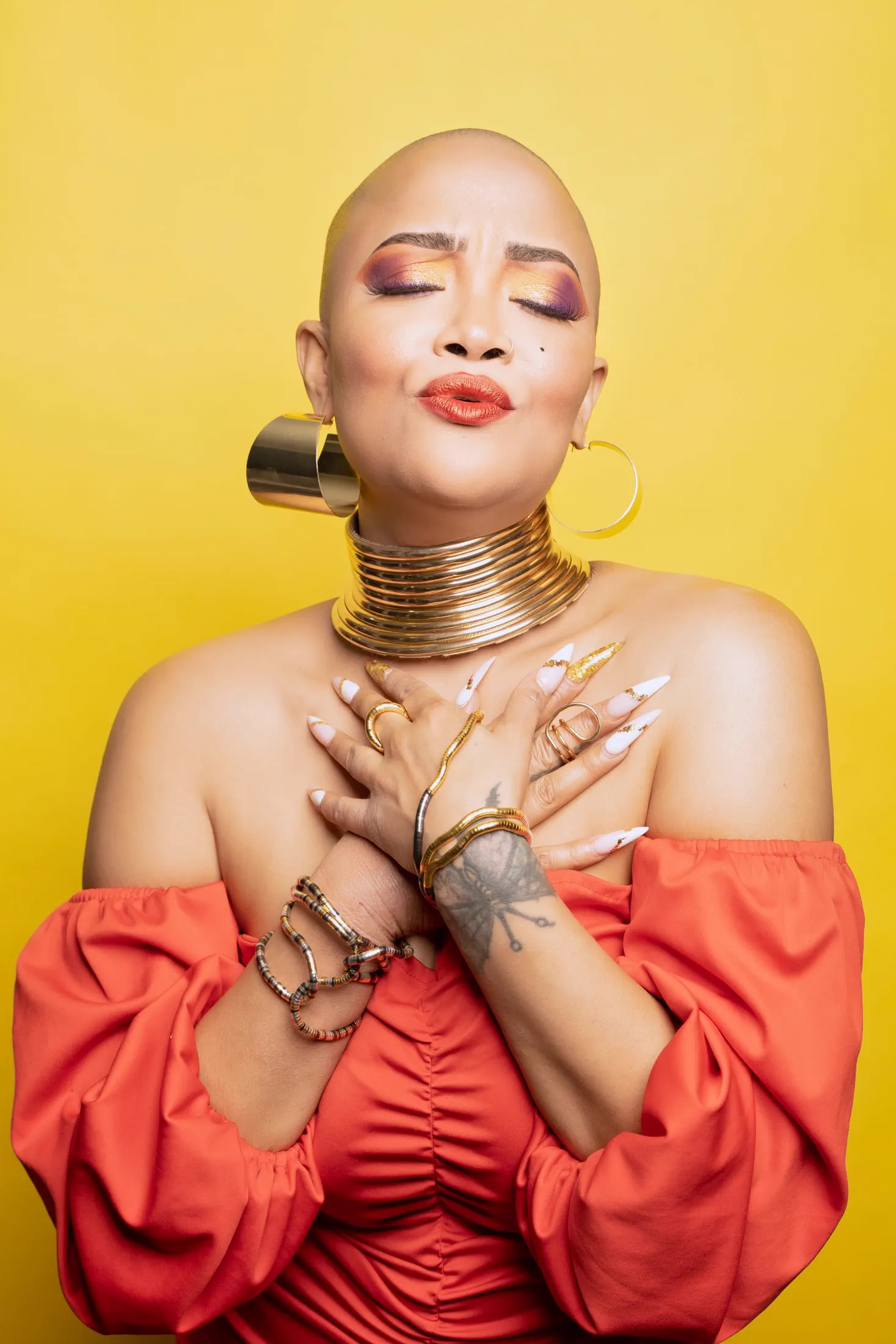 A bald model in a bold red dress poses against a golden-yellow backdrop.
