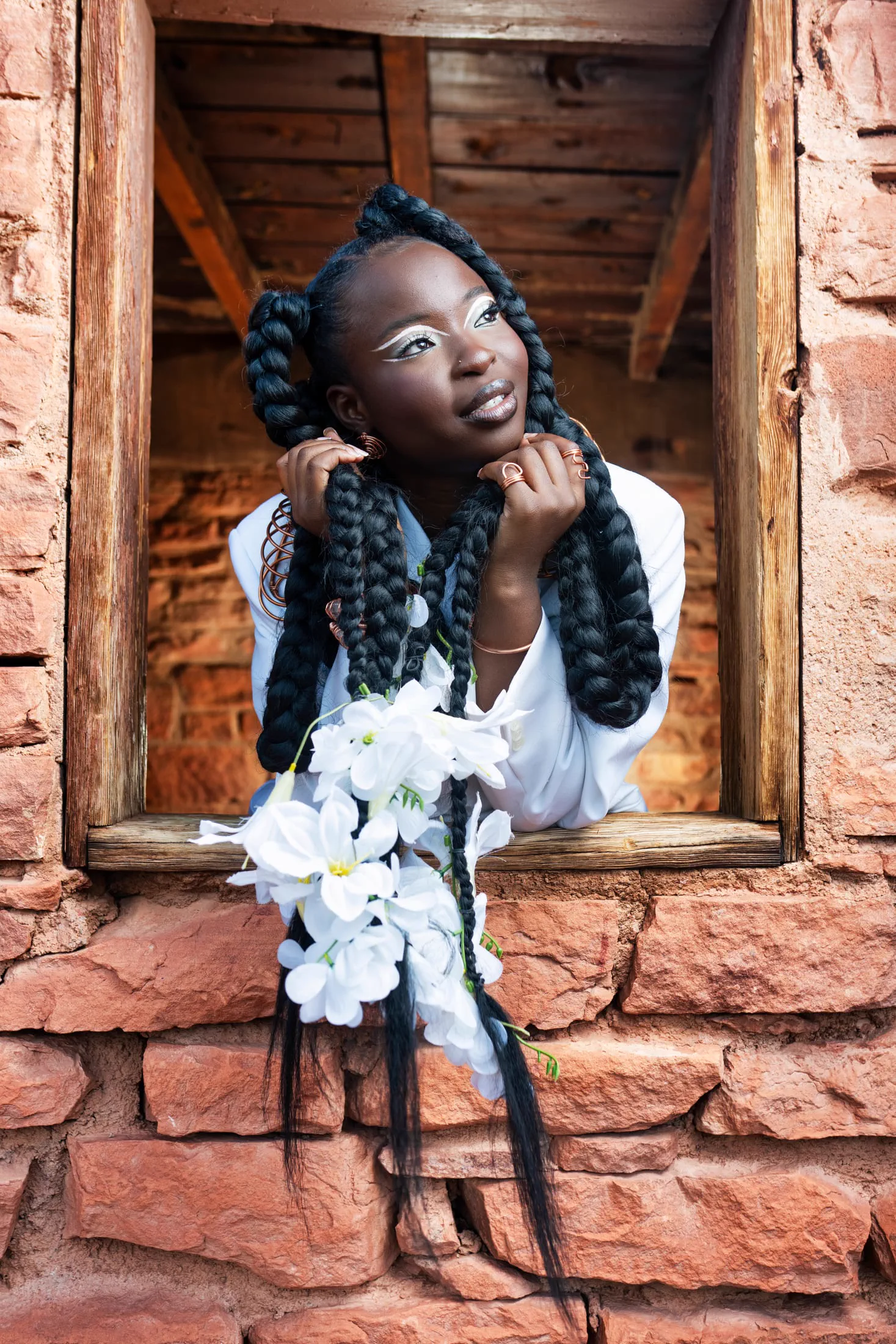 A woman with flower-woven braids looks up thoughtfully from a stone window.