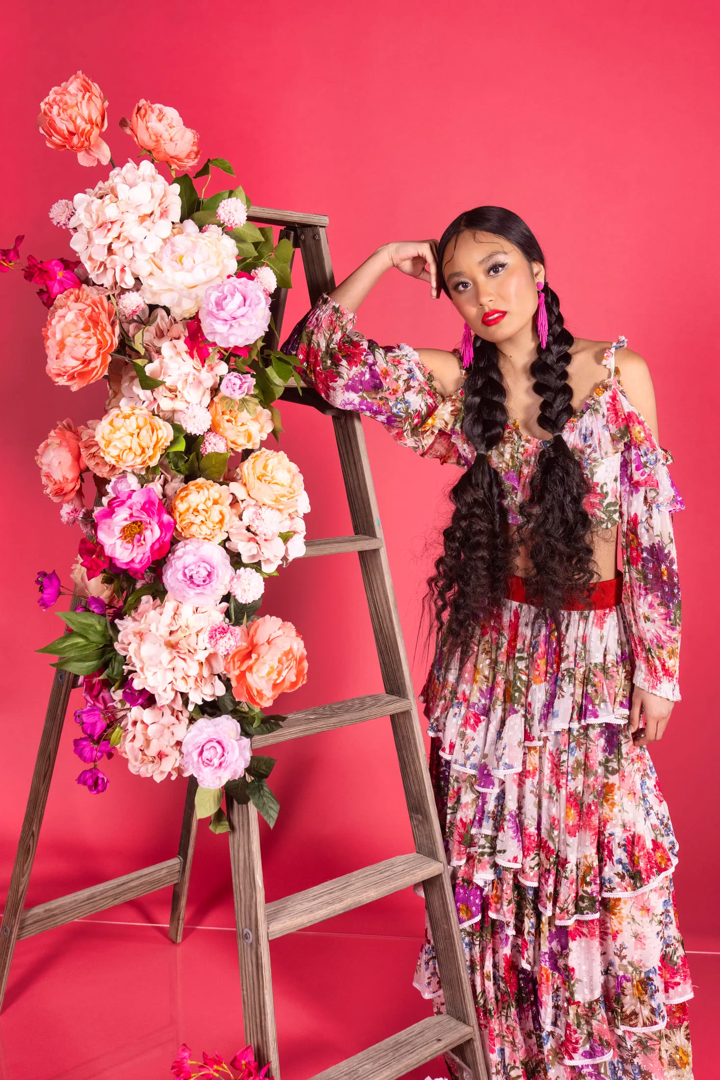 A woman in a floral gown poses beside a ladder styled with pink and coral blooms.