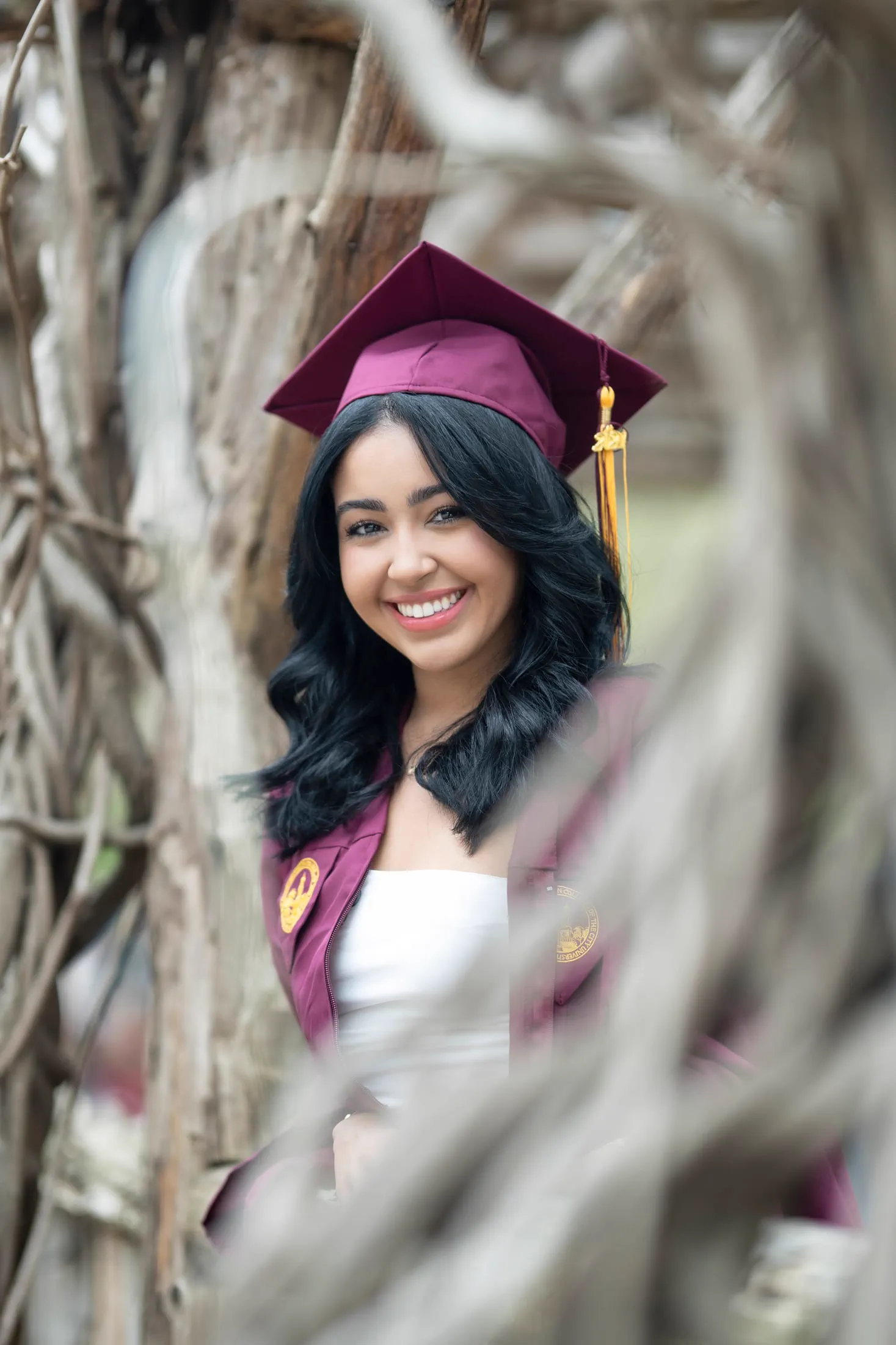 A graduate in cap and gown smiles, framed through bare branches.