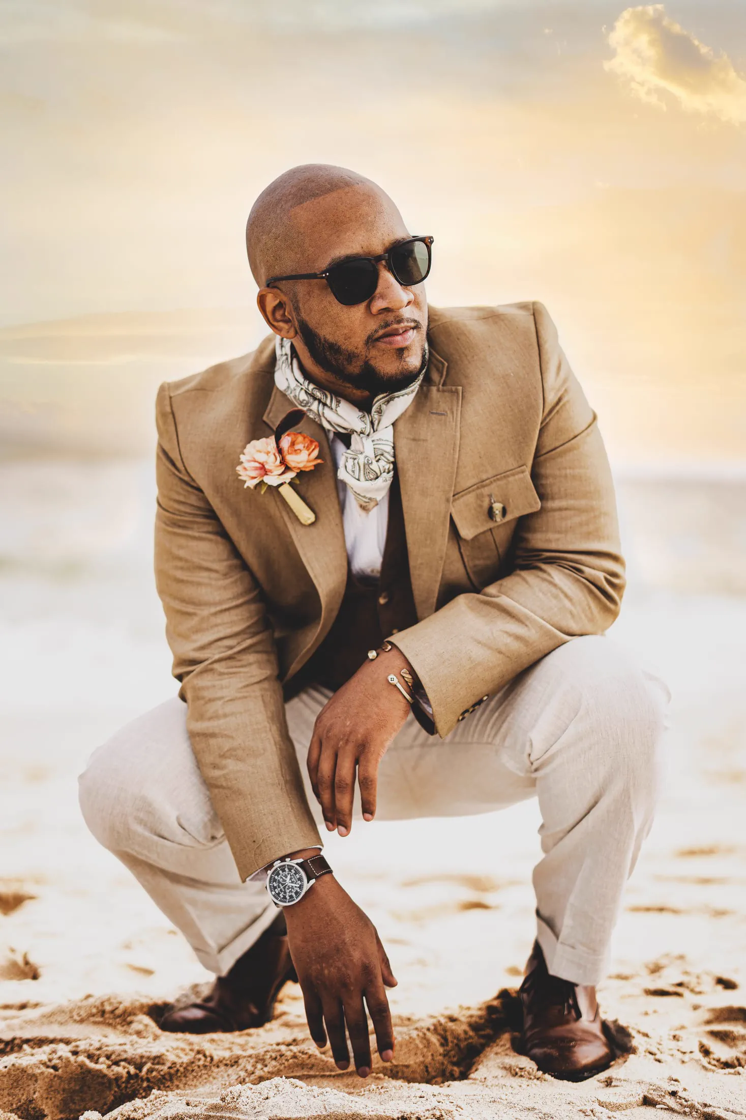 A man in a tan suit and boutonniere crouches on the sand at sunset.