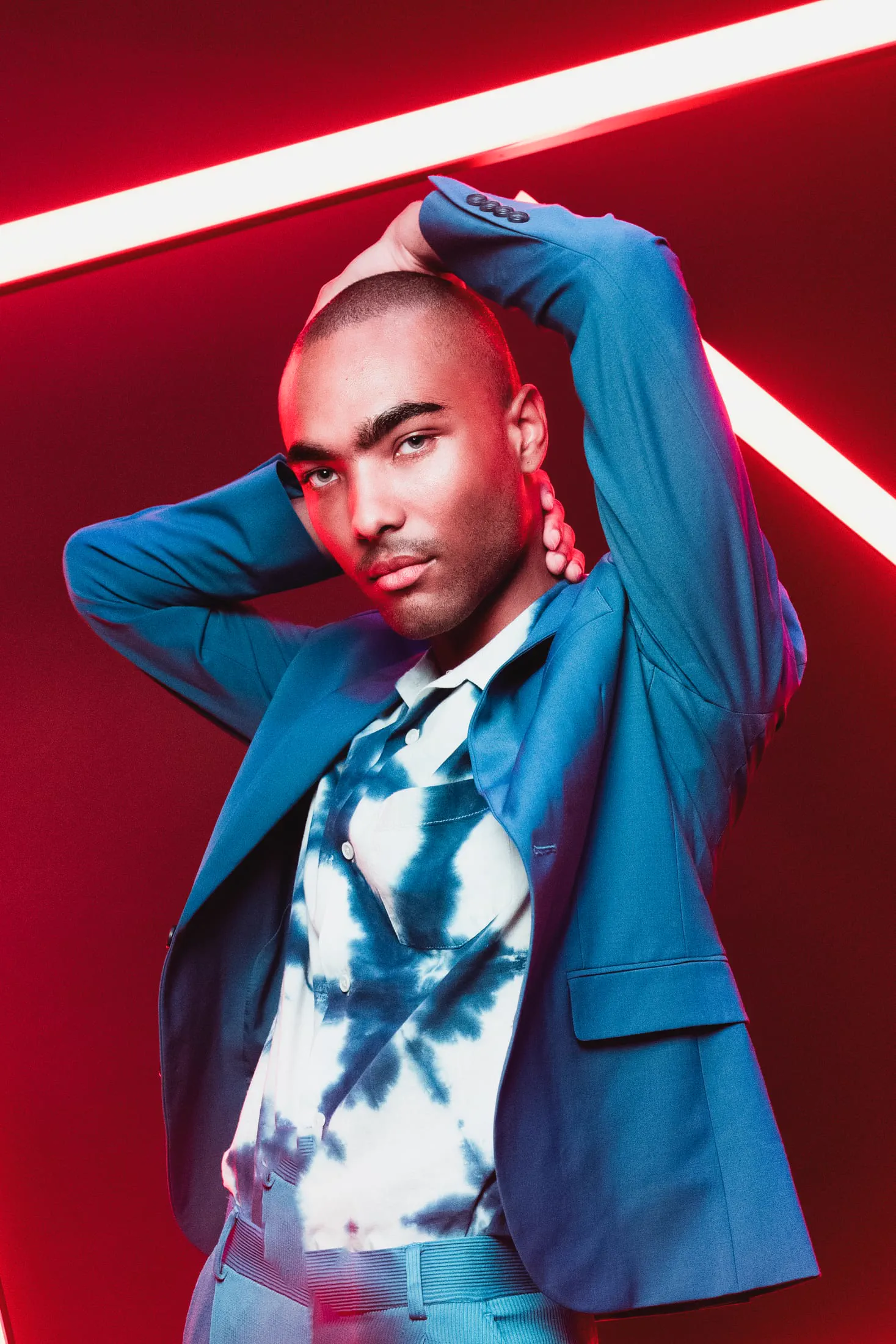 A man in a blue suit and tie-dye shirt poses beneath red neon lines.