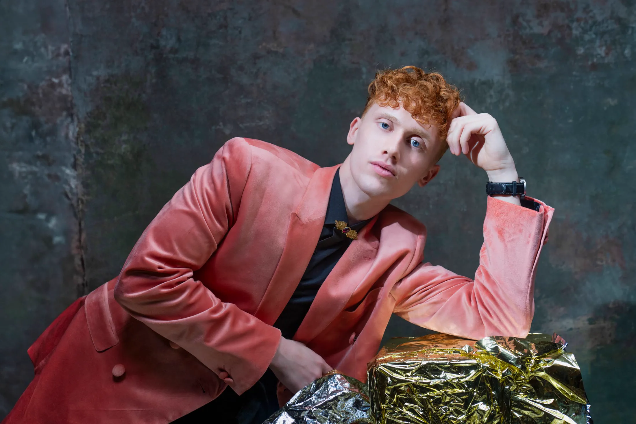 A man in a pink velvet blazer poses against a moody industrial backdrop.