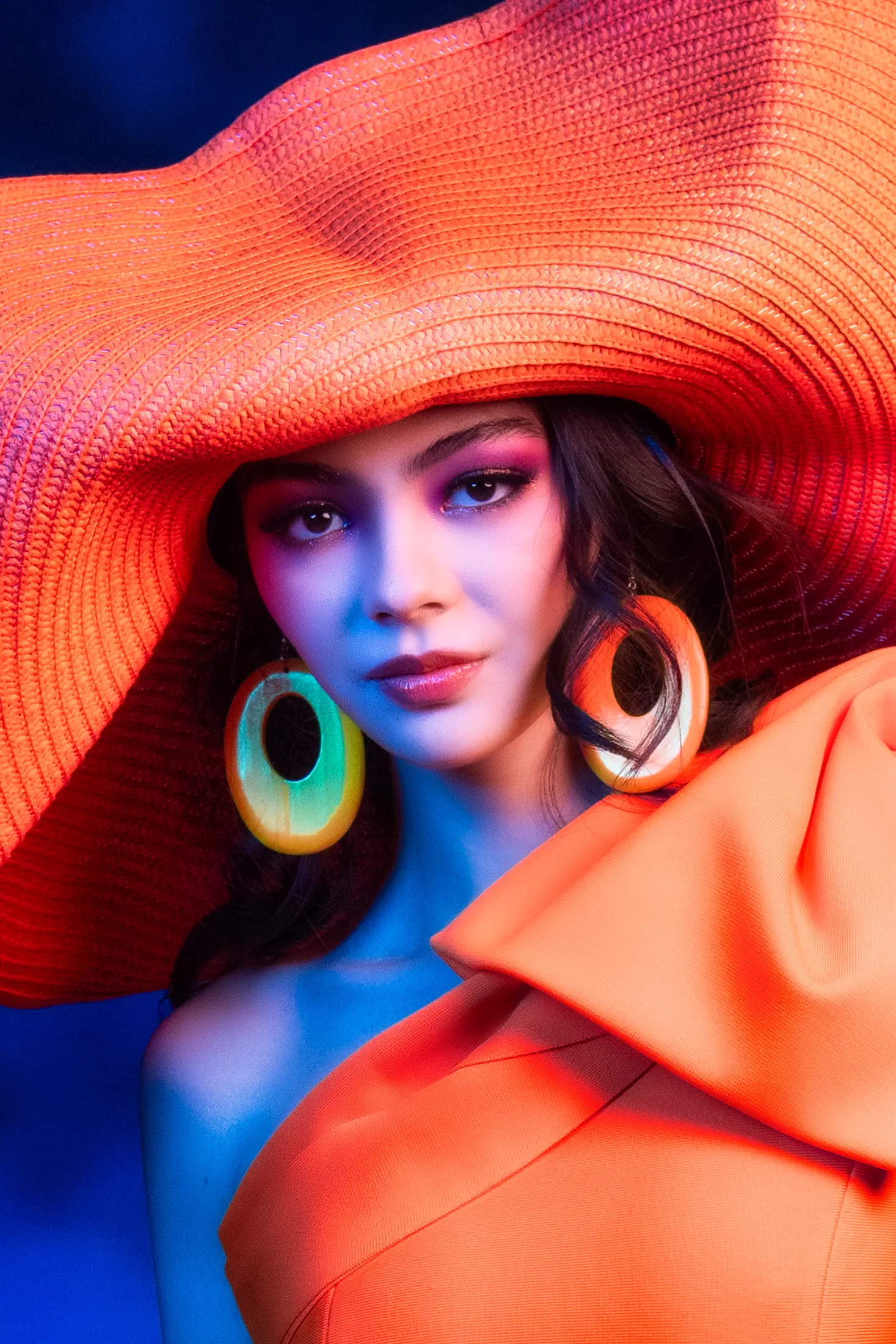 An avant-garde beauty portrait lit in blue and orange, framed by a wide sun hat.