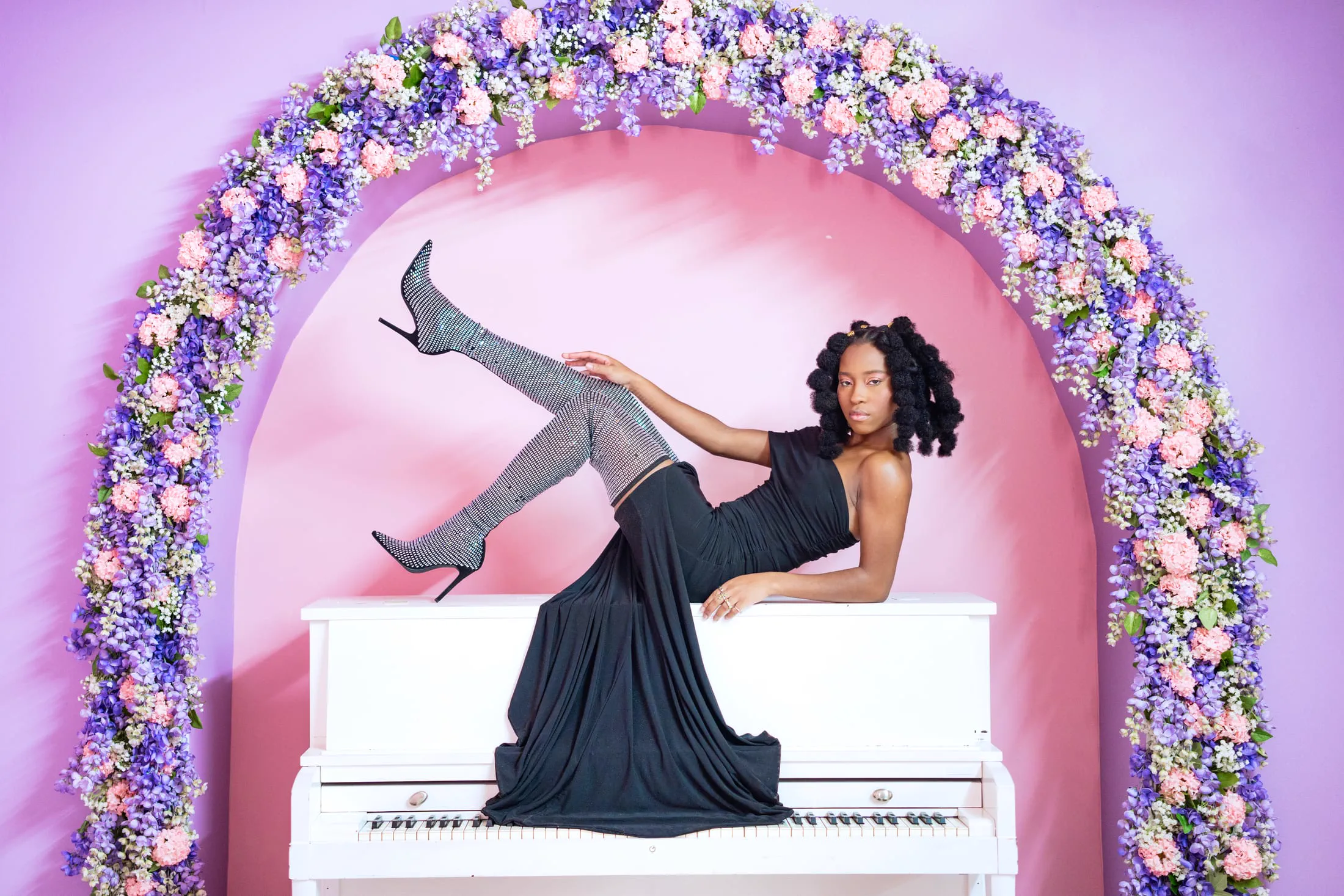 A model reclines atop a white piano beneath a lavender and rose floral arch.