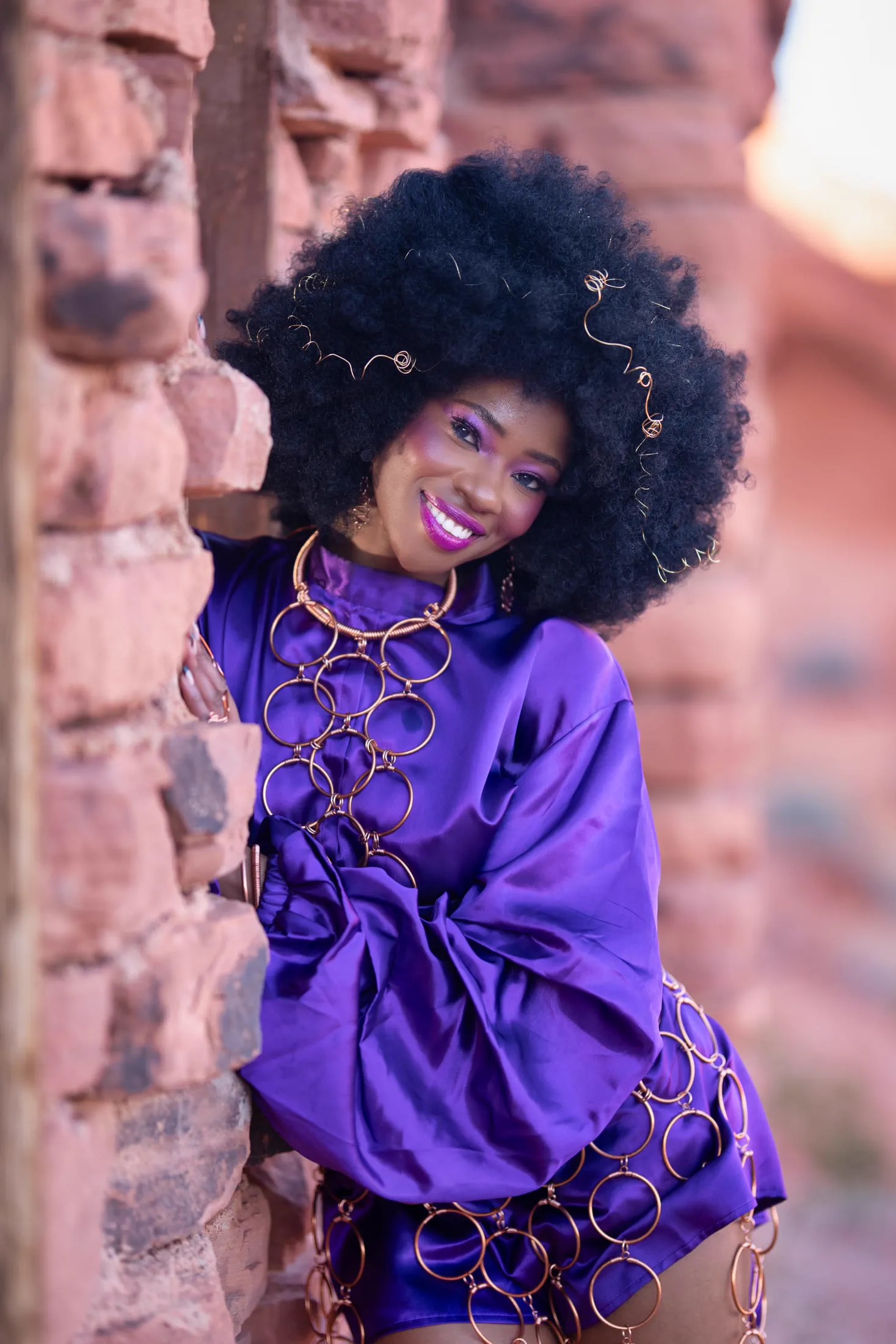 A woman in a purple satin dress and wire-accented afro poses against red rock.