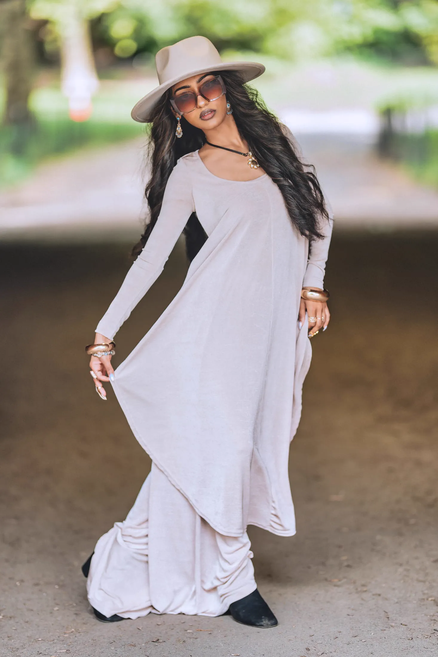 A woman in a tan wide-brim hat and flowing dress poses on a tree-lined path.