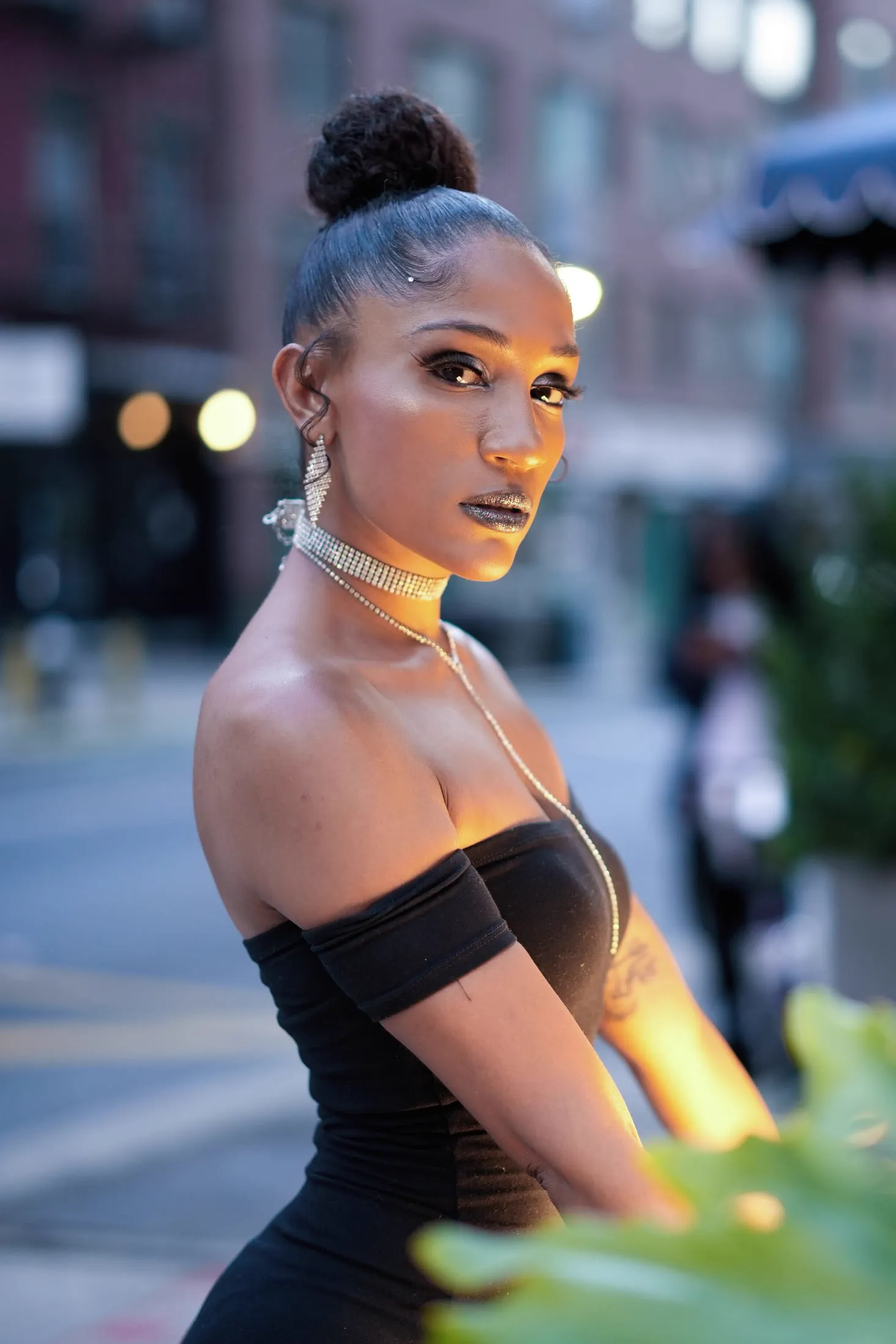 A woman with an updo and silver choker poses on a city street at dusk.