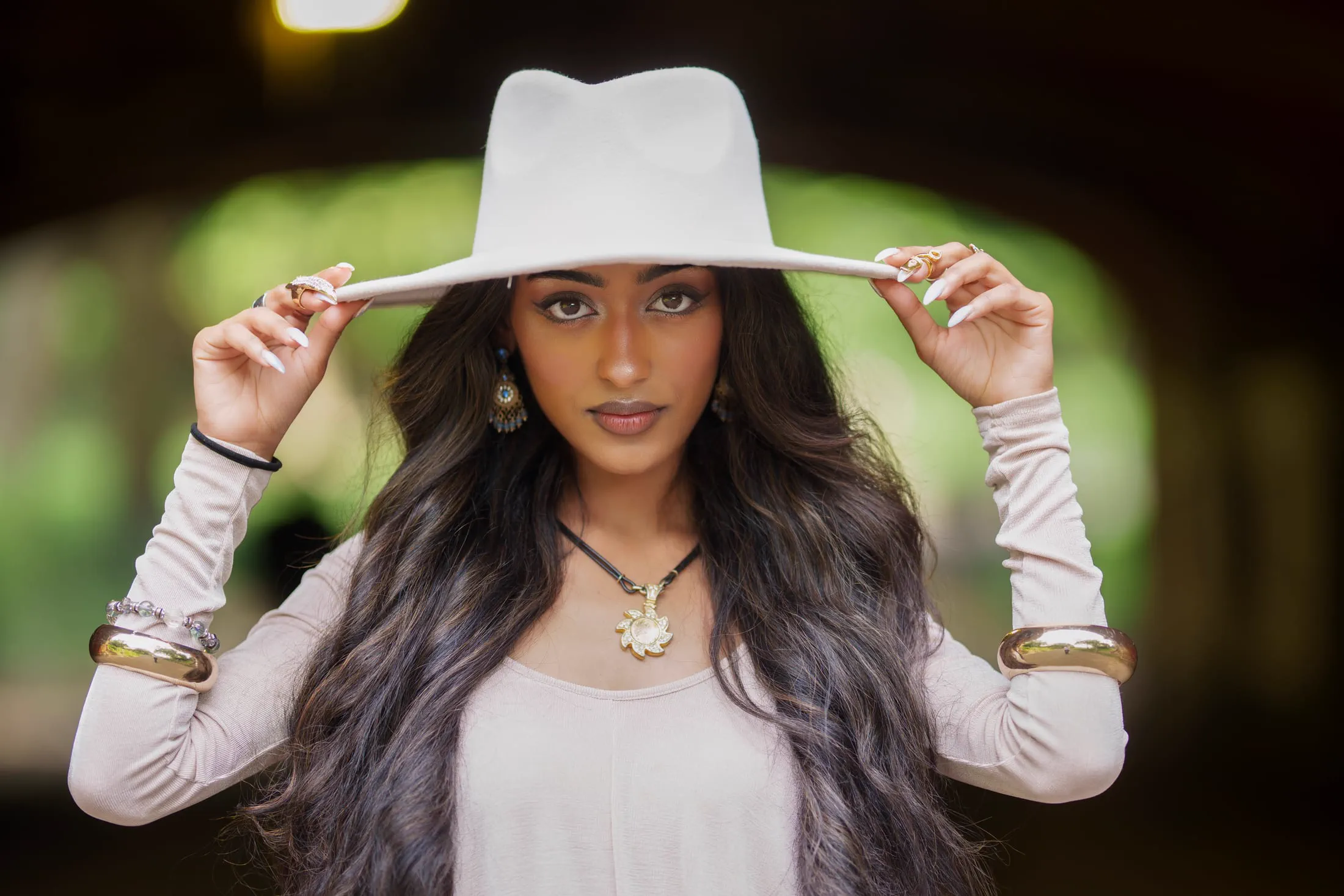 A woman holds the brim of a white hat, jewelry catching soft backlight.