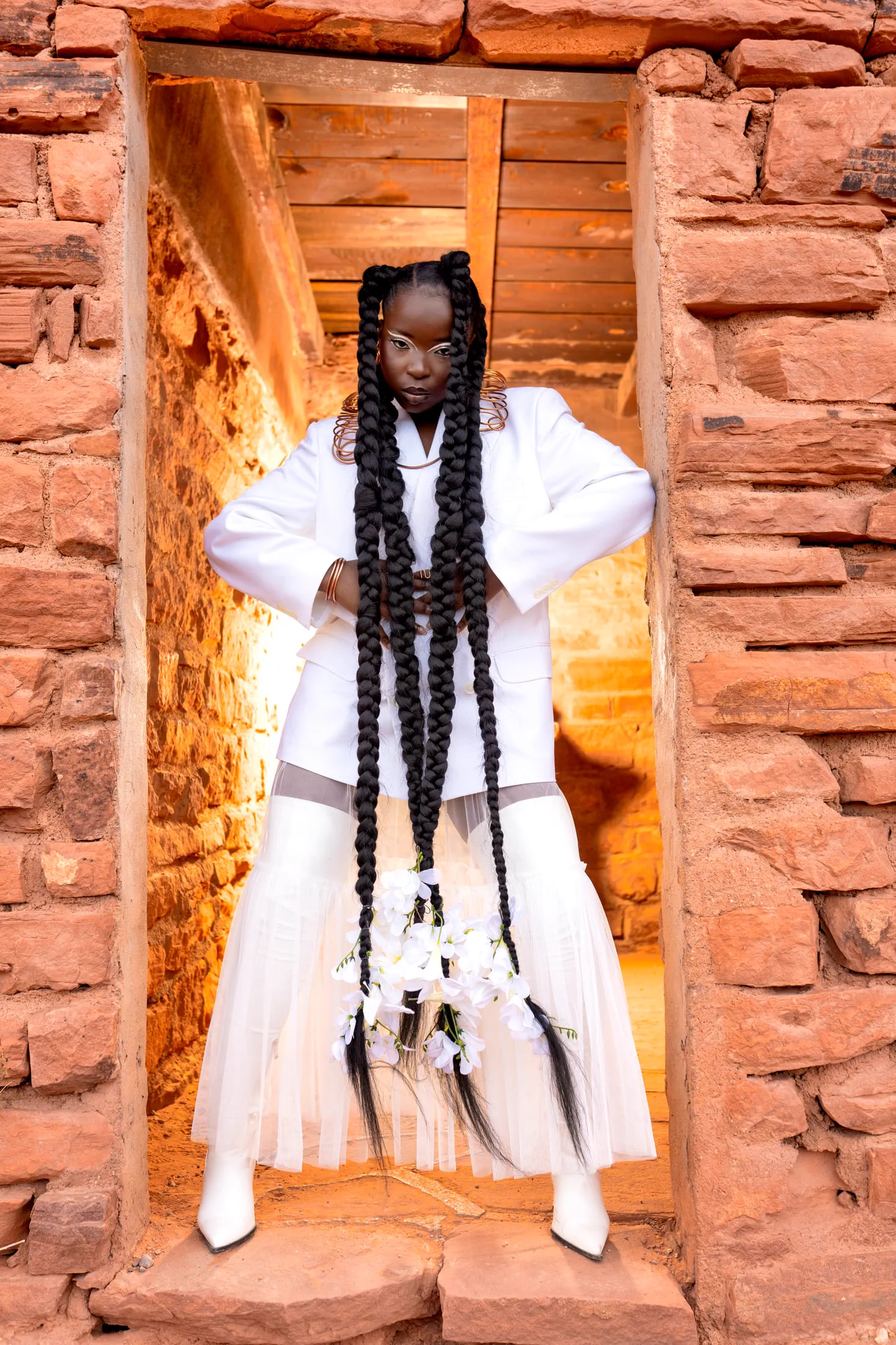 A woman in a white suit and floor-length braids stands in a stone doorway.