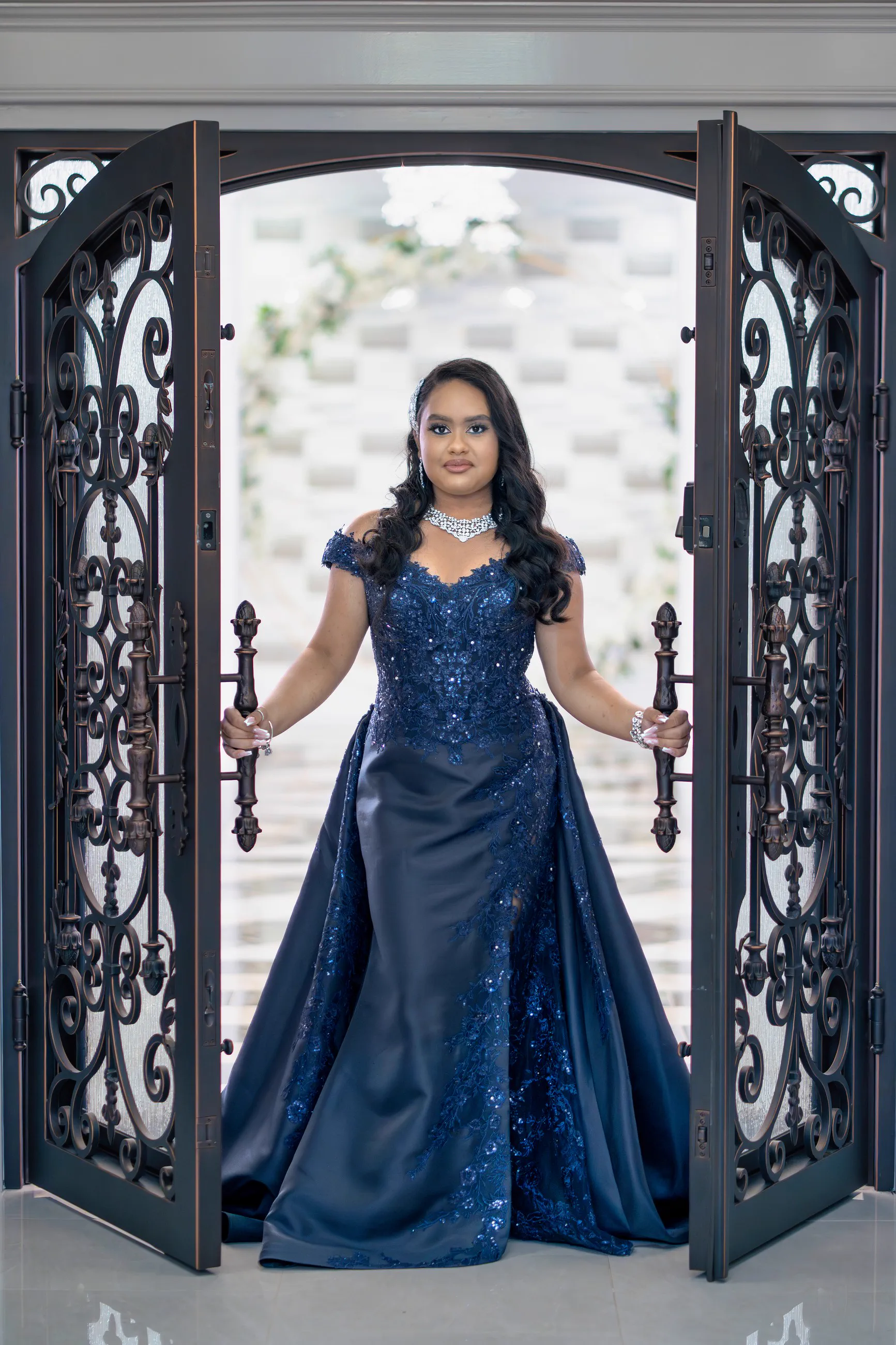 A Sweet 16 celebrant in a deep blue gown stands within ornate iron doors.