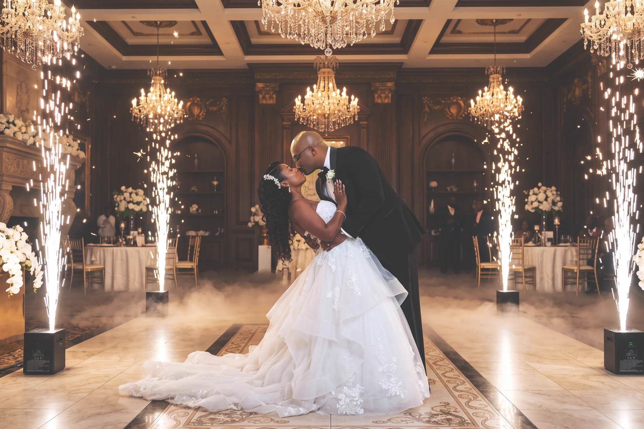 A groom dips his bride for a kiss beneath crystal chandeliers as indoor sparkler fountains light the ballroom.
