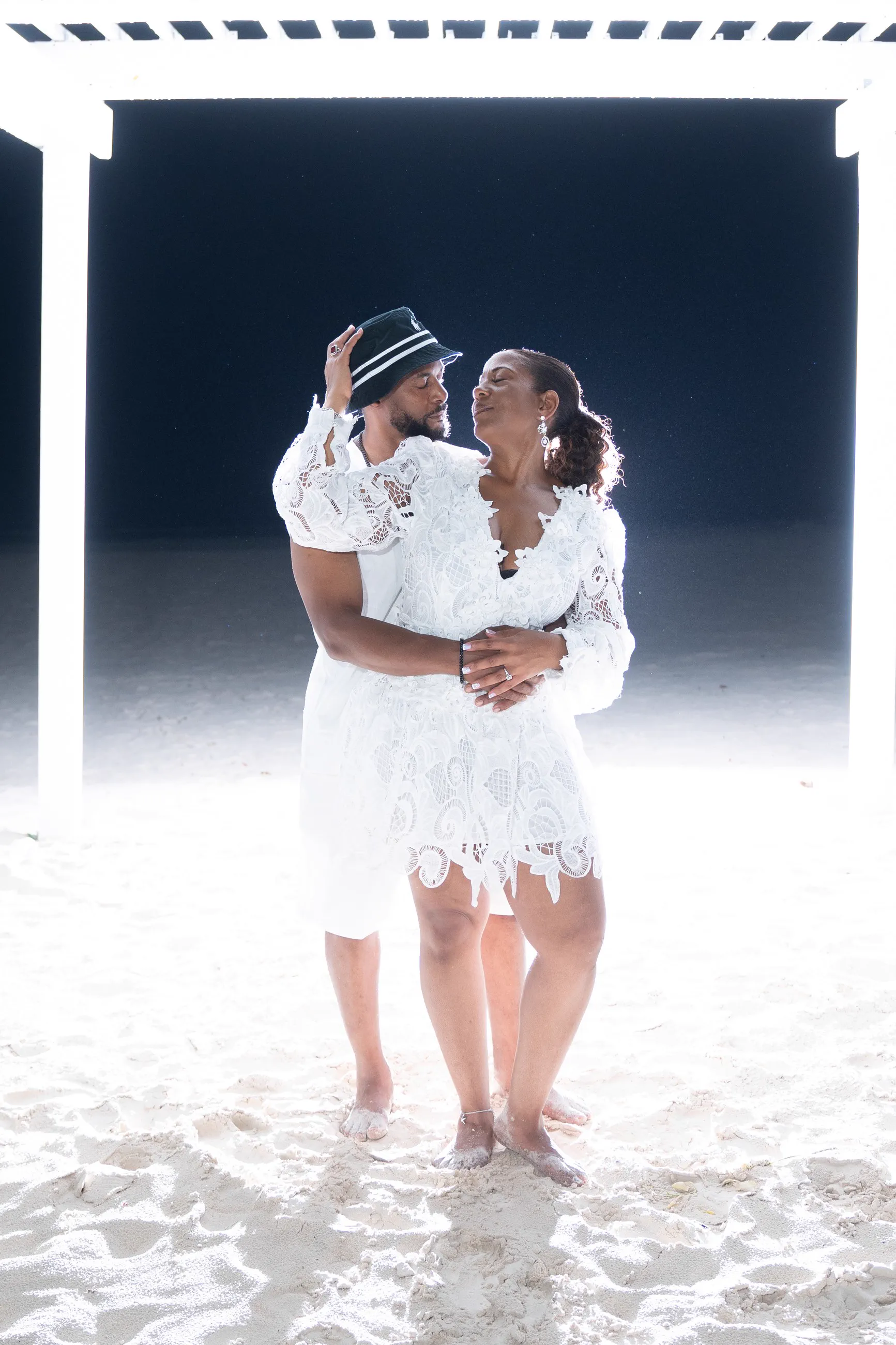 A couple shares a quiet embrace beneath a lit pergola on the sand at night.